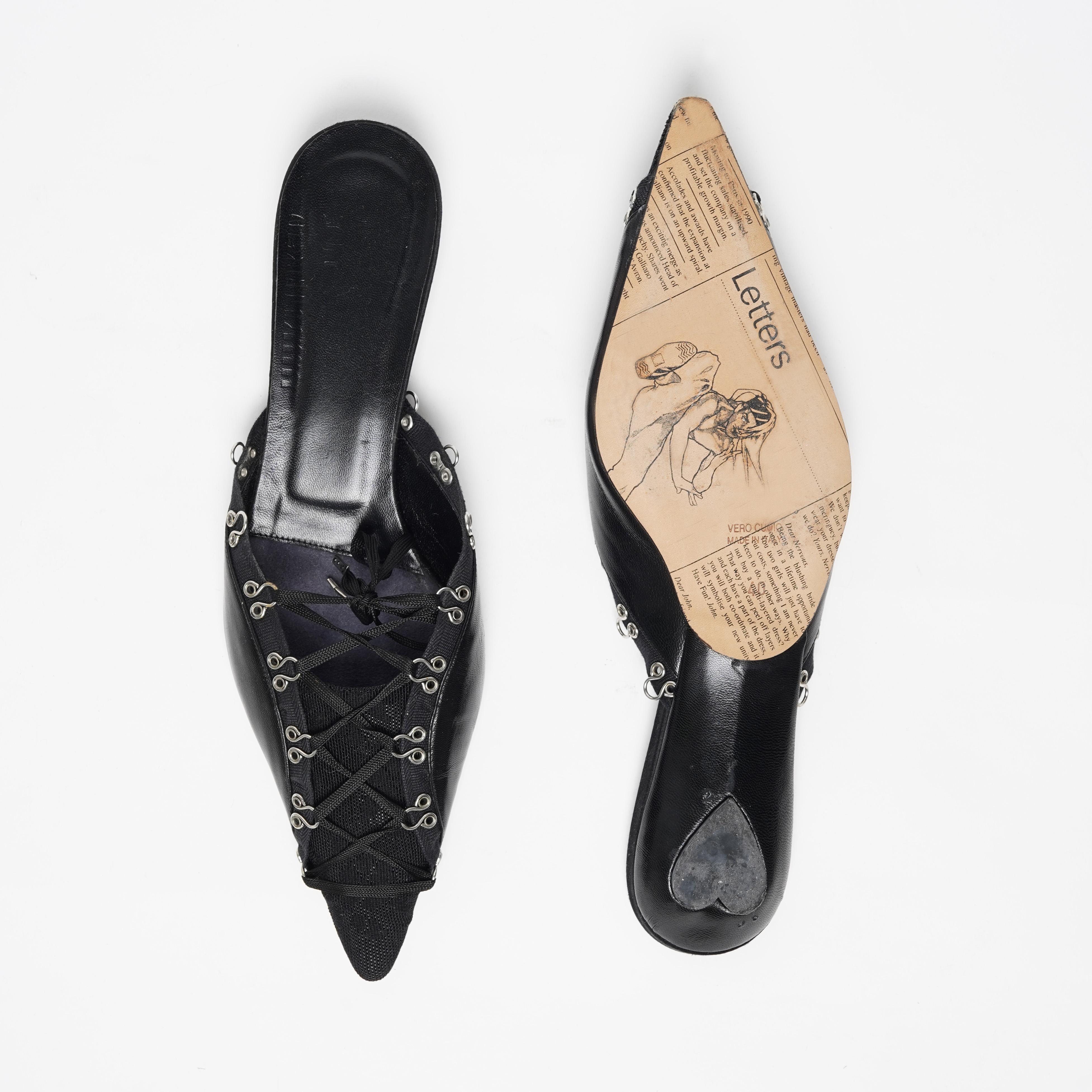 Ti presentiamo un paio di mules corsetto lingerie in pelle nera di John Galliano con suola stampata a gazzetta e tacco a forma di cuore. Queste mules allacciate con occhielli di John Galliano sono davvero au courant. Presentano l'iconoclasta stampa