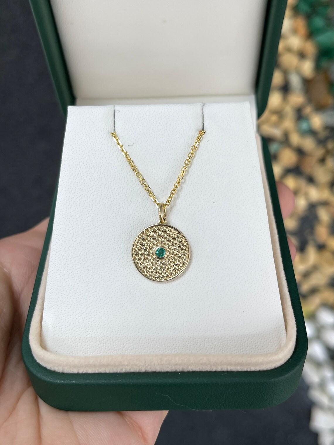 Questo splendido ciondolo a forma di smeraldo d'oro ha la forma di un cerchio con un'accattivante finitura strutturata sulla superficie d'oro. Al centro del cerchio, uno smeraldo minuscolo di taglio rotondo è incastonato in un castone e mostra un