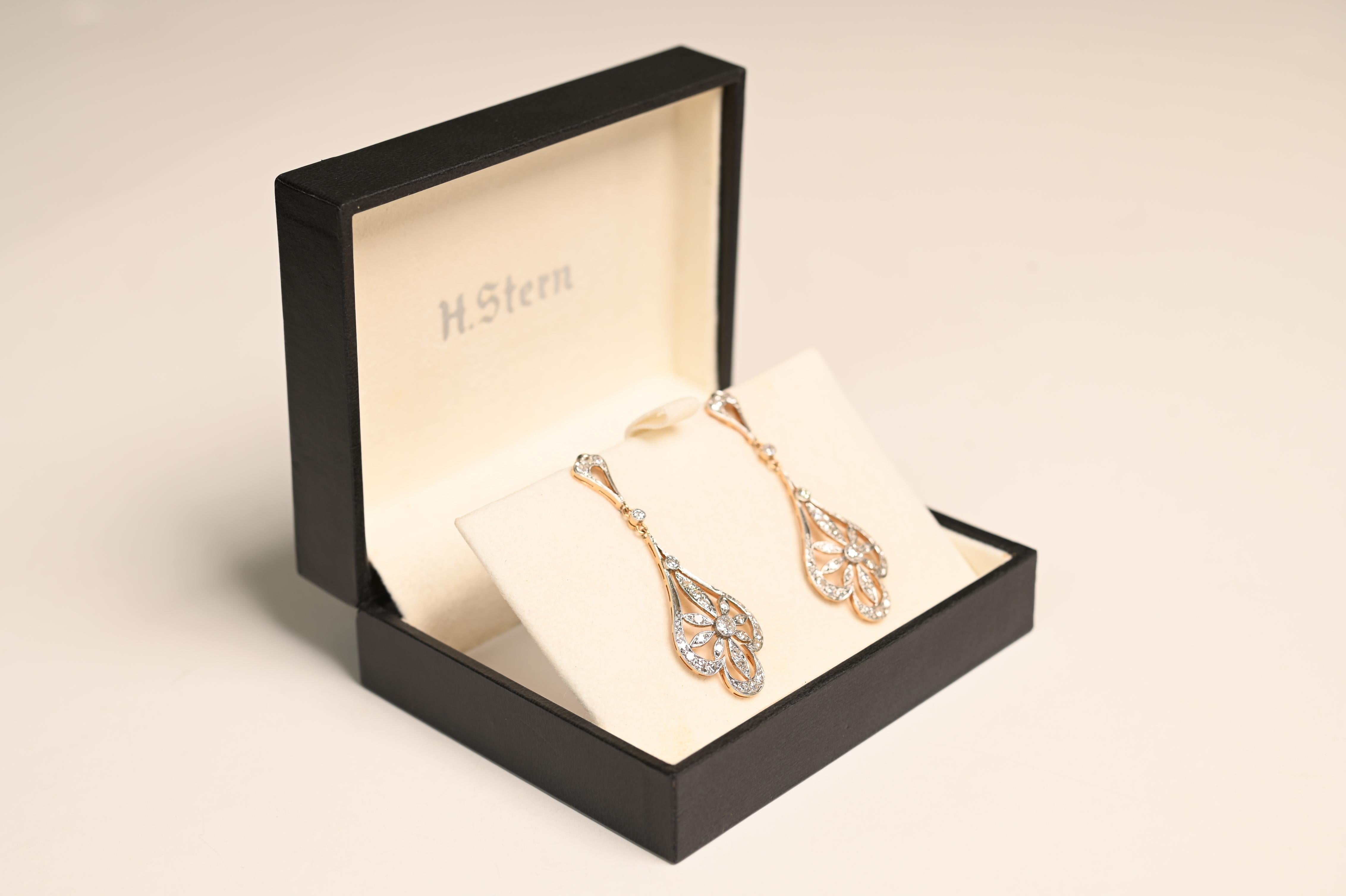 These elegant Edwardian-style dangling earrings feature a timeless design with two bezel-set transitional cut diamonds, each weighing approximately 0.10-carats. The center stones are beautifully surrounded by a delicate floral motif, accented by