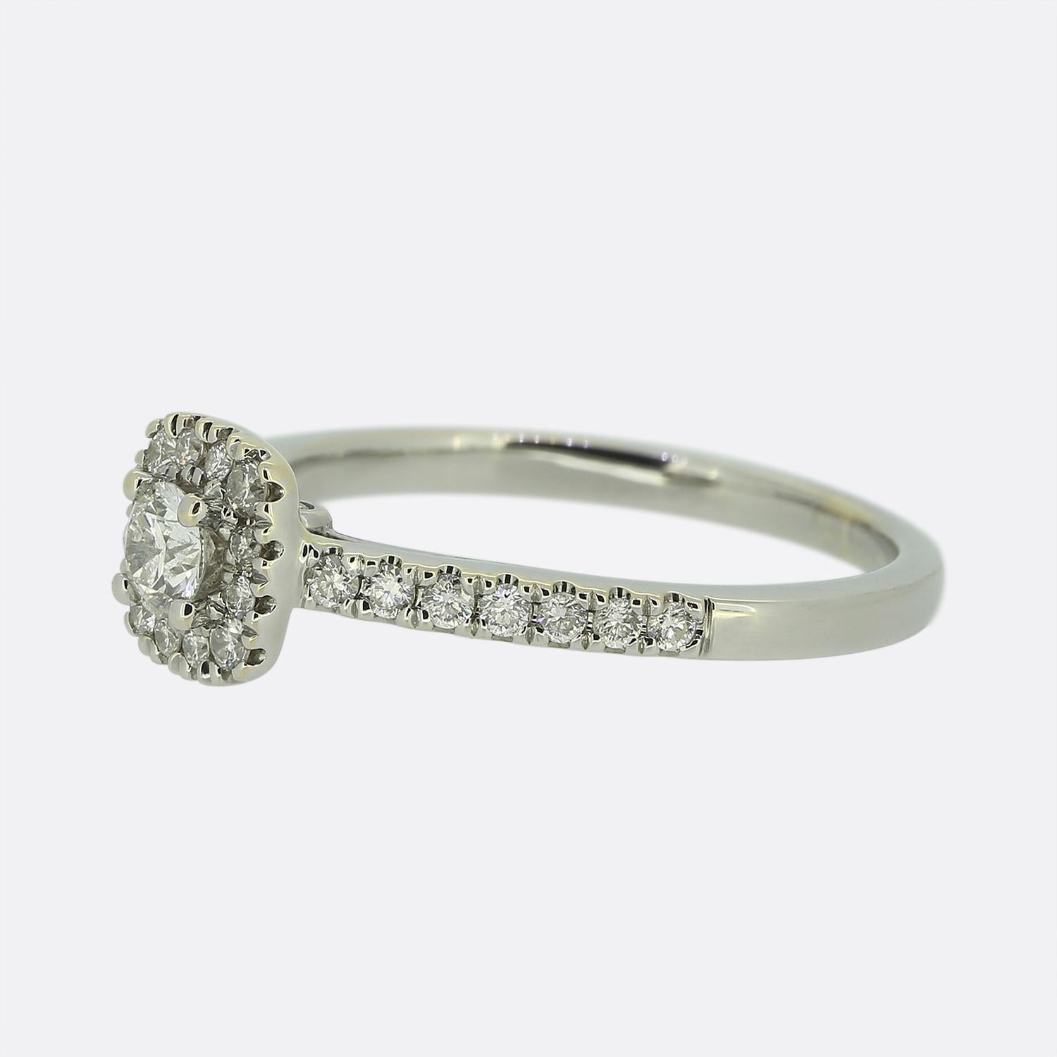 Qui abbiamo un anello di fidanzamento con diamante in oro bianco da 18 carati. Il diamante centrale è un taglio brillante rotondo da 0,20 carati incastonato in una montatura a quattro griffe. Questa pietra principale si trova su uno sfondo composto