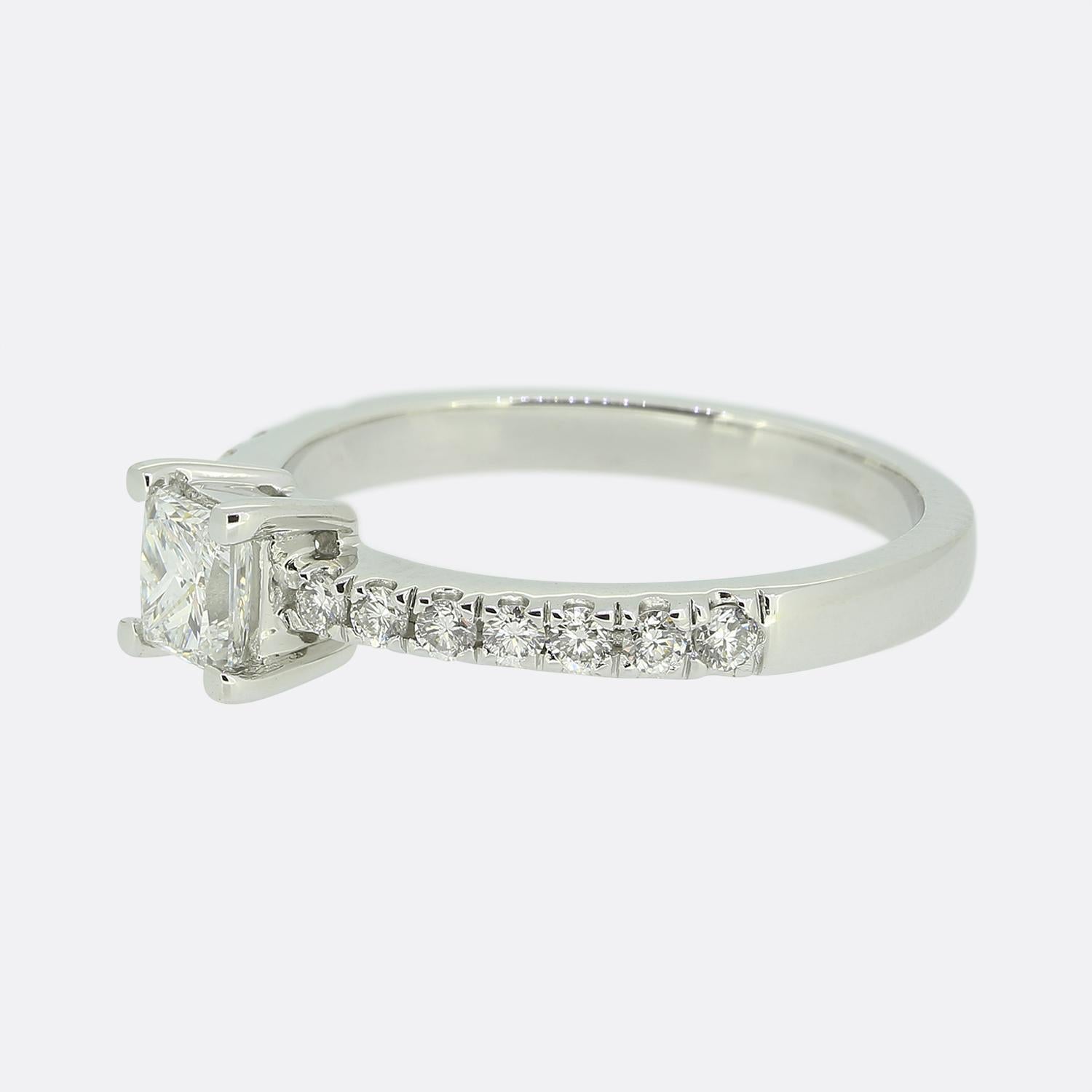 Hier haben wir einen Solitärring aus 18 Karat Weißgold mit einem Diamanten im Prinzessschliff. Der zentrale Diamant im Prinzessinnenschliff ist in einer Fassung mit 4 Krallen gefasst. 7 runde Diamanten im Brillantschliff führen in einer einzigen
