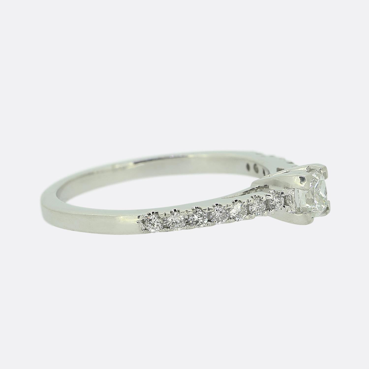 Here we have an 9ct white gold diamond engagement ring. The focal princess cut diamond sits risen at the centre of the face whilst being flanked on either should by a single row of seven round brilliant cut diamonds whilst the back of the shank