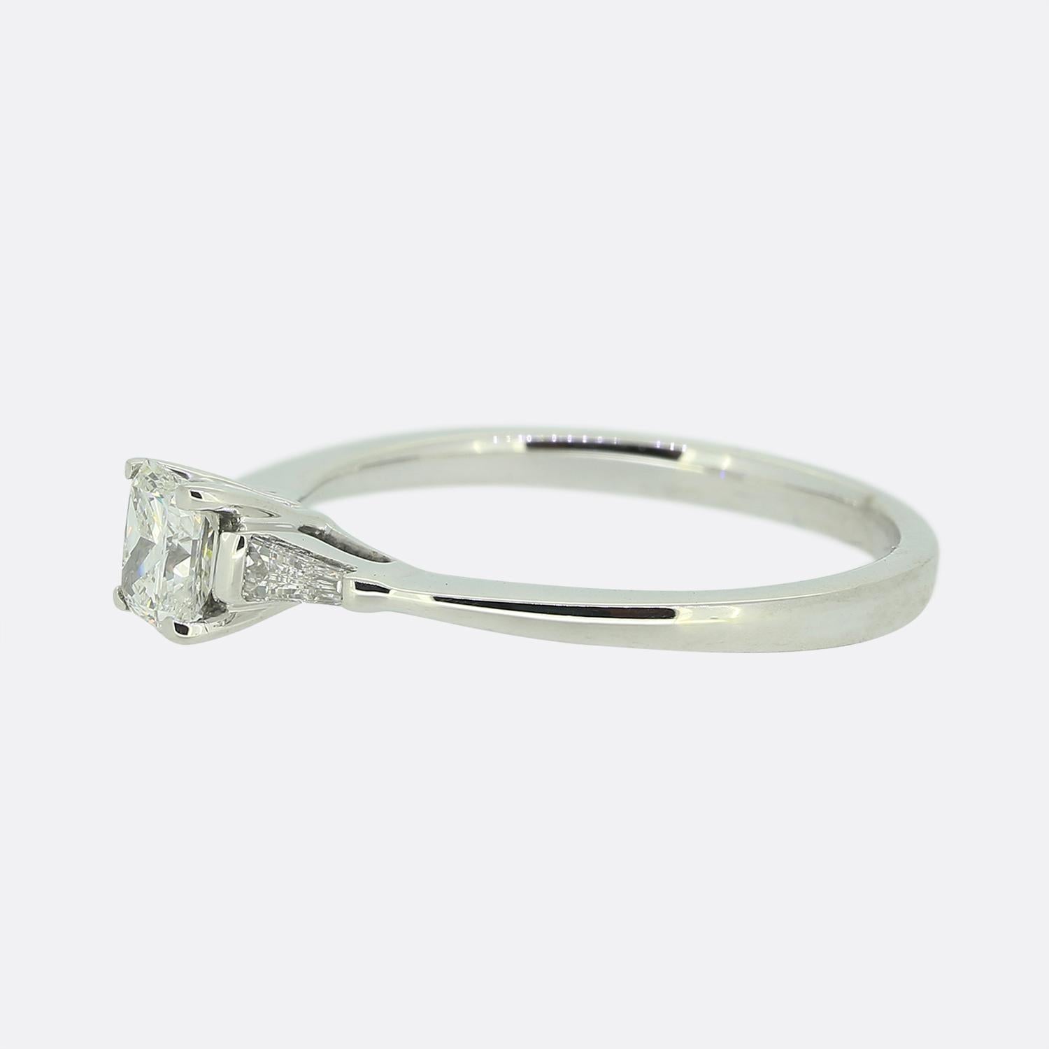 Hier haben wir einen Ring aus 18 Karat Weißgold mit mehreren Diamanten. In der Mitte befindet sich ein Diamant im Prinzessinnenschliff, der in einer erhöhten Fassung mit vier Krallen gefasst ist, mit einem schmalen, konischen Baguette-Diamanten auf