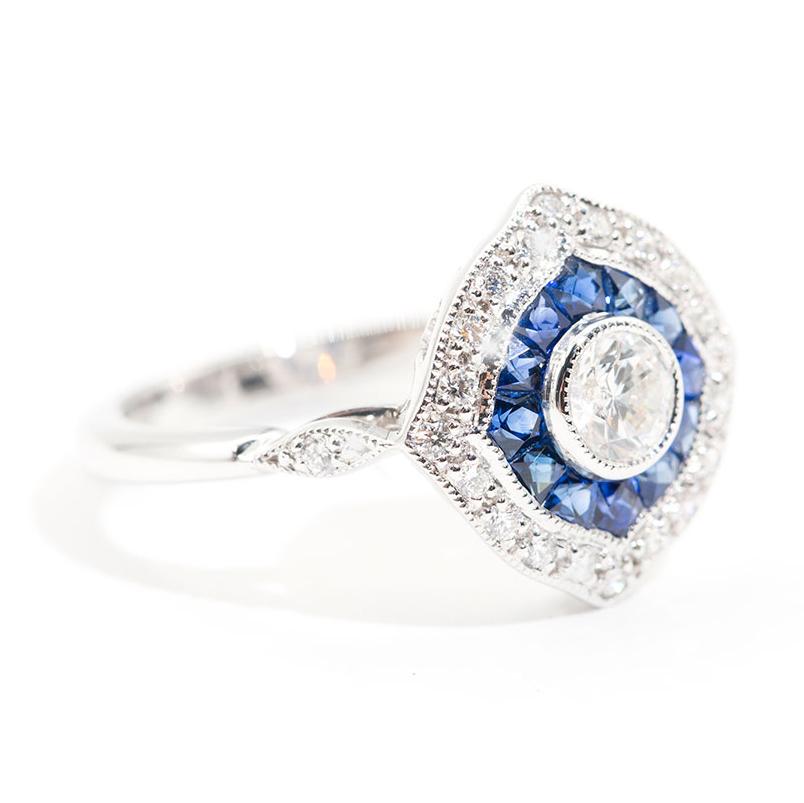 Forged in 18 carat white gold is this art deco inspired ring featuring a central 0.37 carat round brilliant cut diamond encompassed with a border of bright deep blue colour custom cut natural sapphires and sparkling round brilliant cut diamonds. We