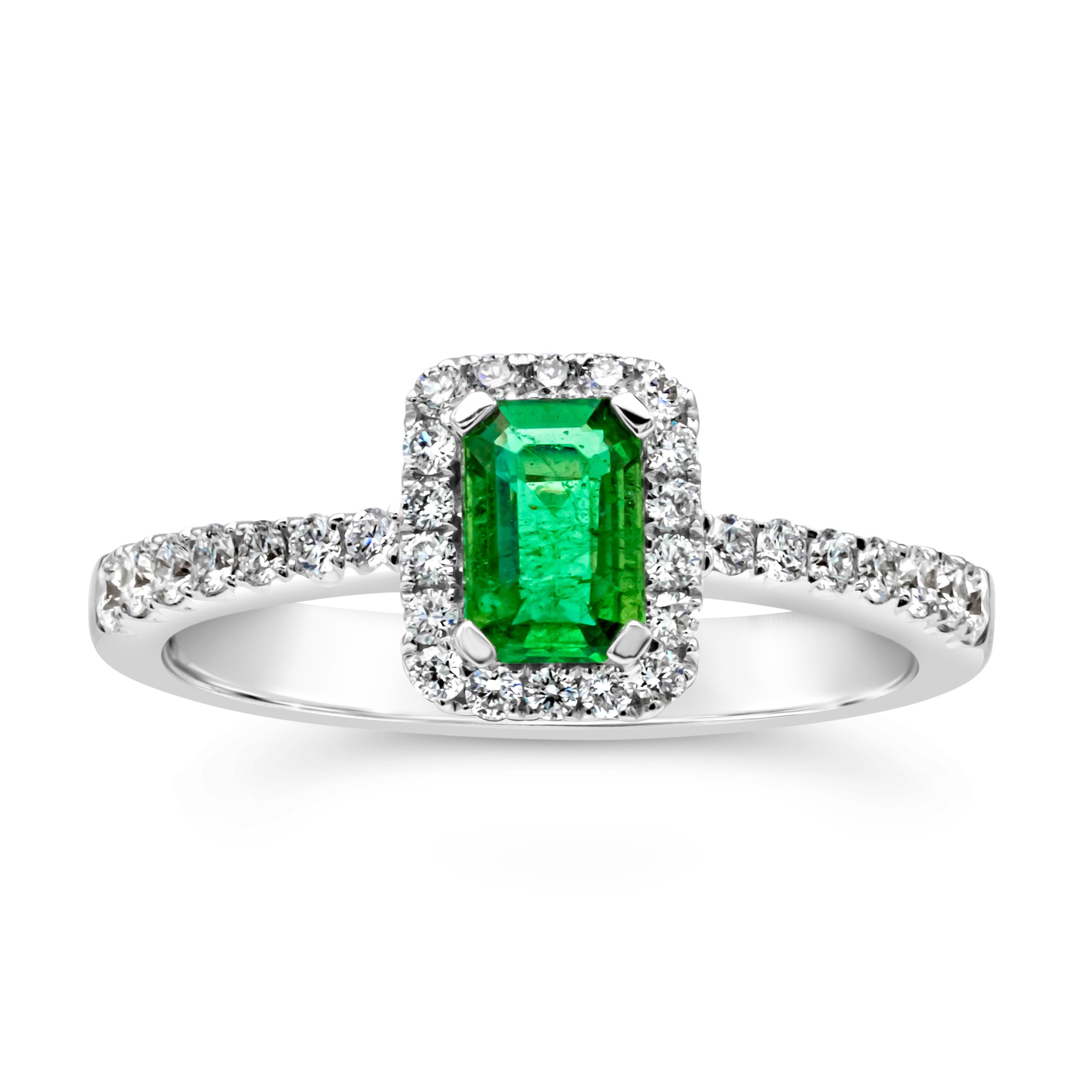 An appealing halo engagement ring style showcasing an emerald cut green emerald weighing 0.49 carats total, set in a four prong basket setting. Elegantly surrounded by a row of brilliant round diamonds that continues on to the shank of the ring