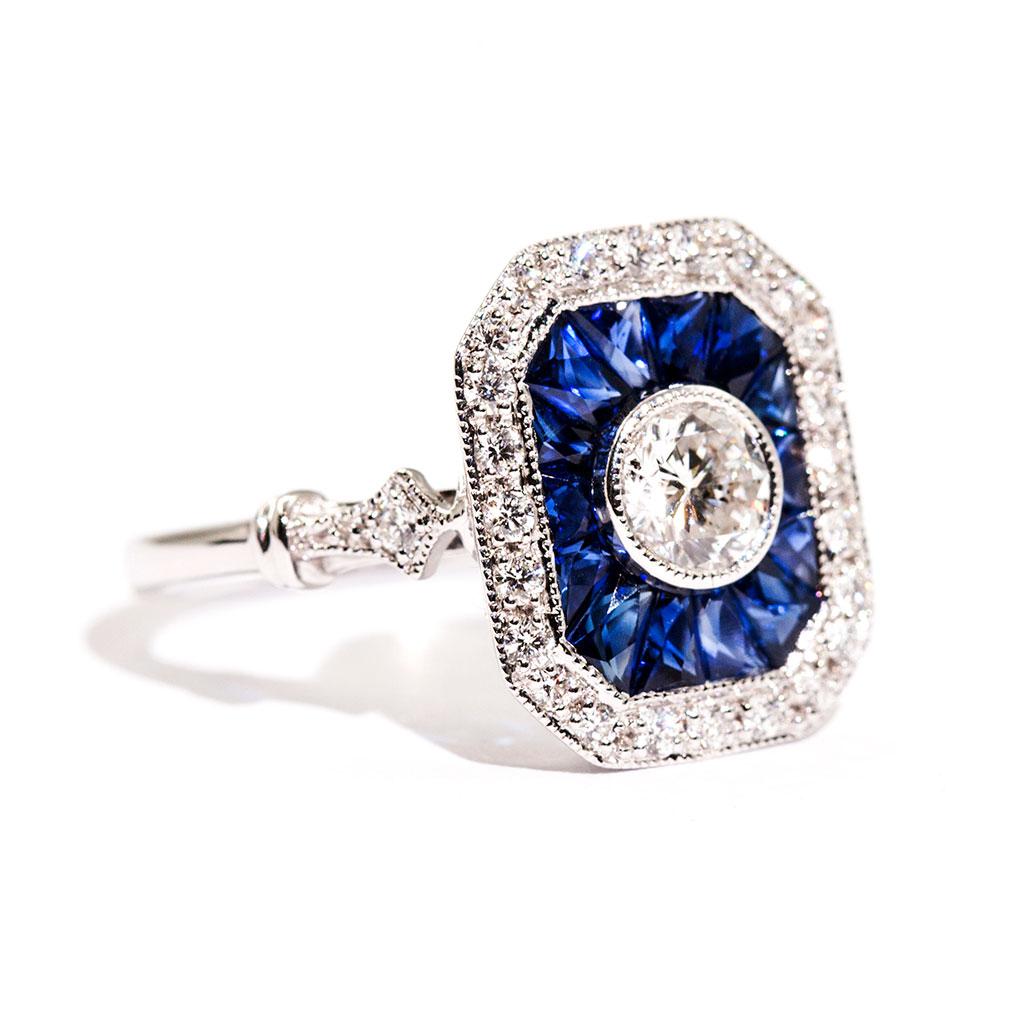 Forged in 18 carat white gold is this vintage inspired ring that features a central certified 0.50 carat round brilliant cut diamond that is encompassed with a border of bright deep blue colour custom cut natural sapphires totalling 0.40 carats and