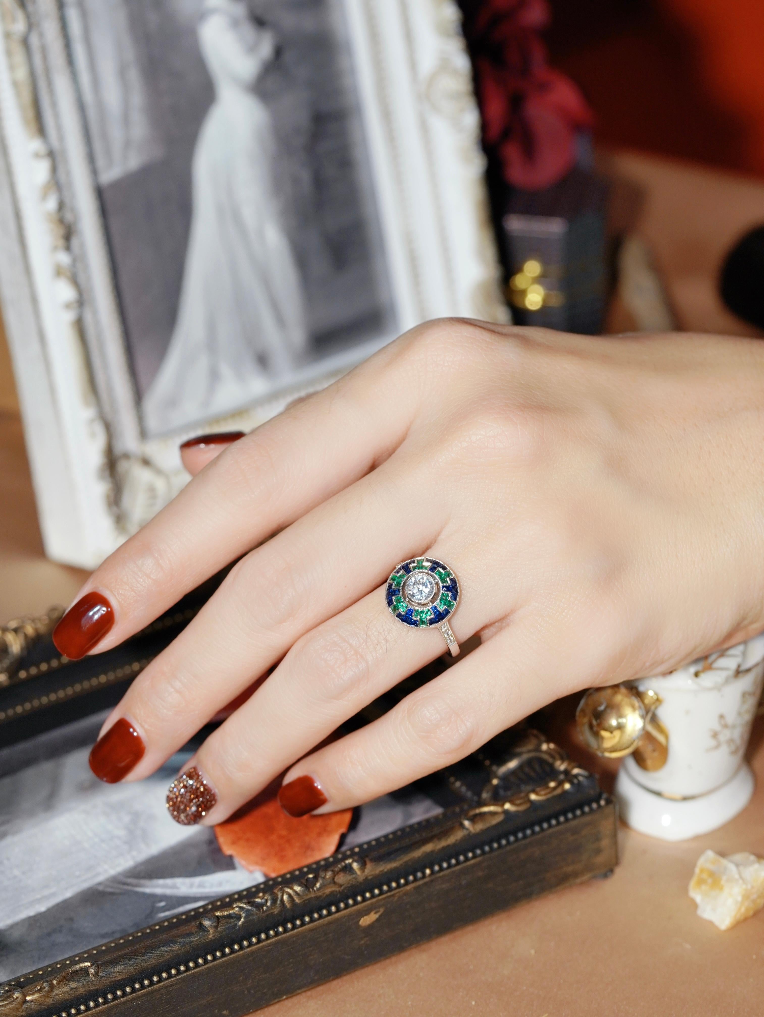 Un elegante anello con diamante, zaffiro blu e smeraldo. Il diamante rotondo centrale è incastonato in modo sicuro nell'oro bianco come centro di questo splendido anello. Disegnati per assomigliare a un'elegante ruota geometrica, zaffiri blu e