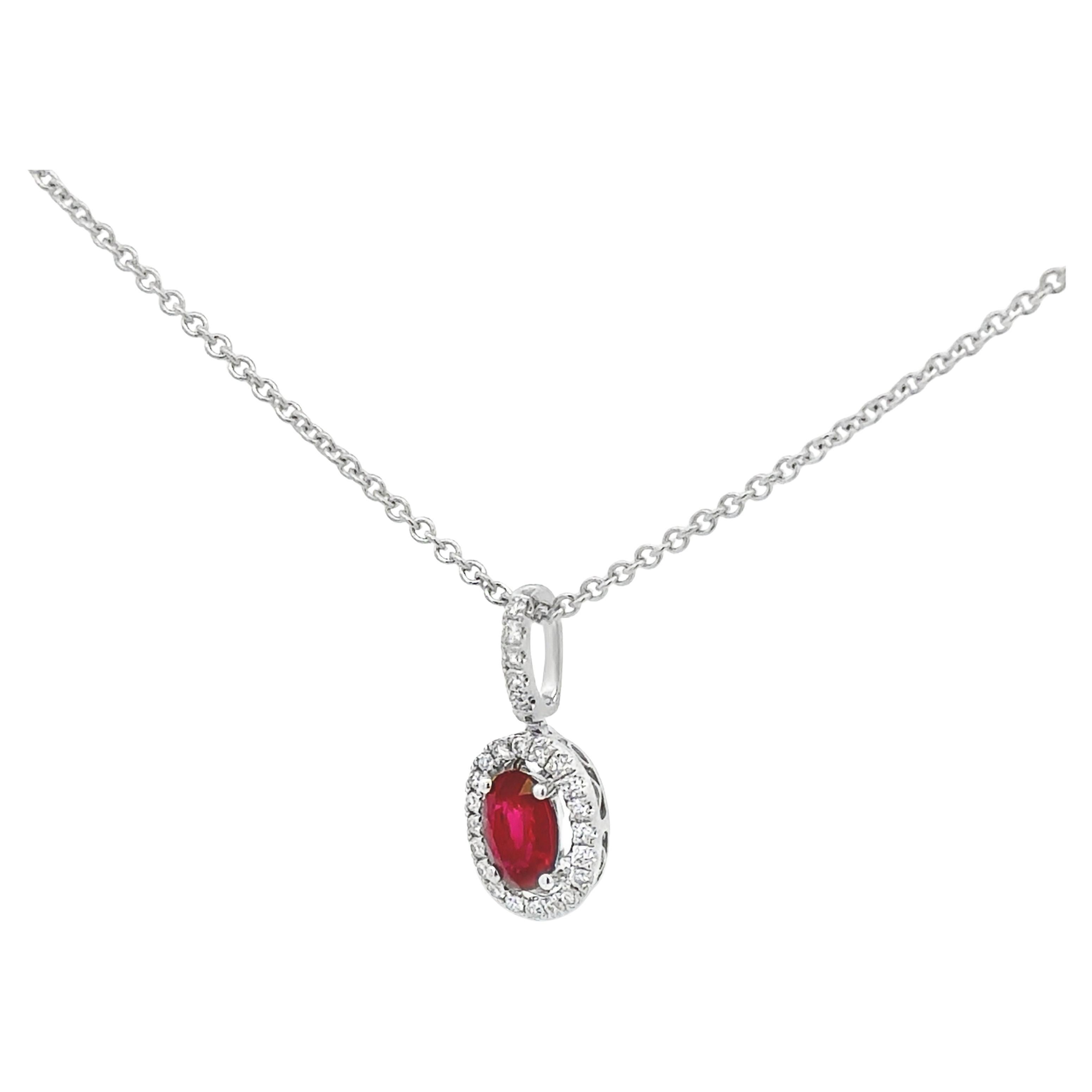 A stylish pendant necklace, featuring a 0.56 carat oval cut ruby, set on four rounded prongs. Accented by a halo of round cut diamonds, set on an open-work design and a diamond encrusted bail. Accent diamonds weigh 0.13 carat total. Finely made with