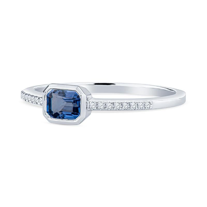 This dainty ring features a 0.58 emerald cut natural blue sapphire set east-west accented by 0.04 carat total weight in round diamonds along the band set in 14 karat white gold. This ring is a size 6.5 but can resized upon request. Wear alone or