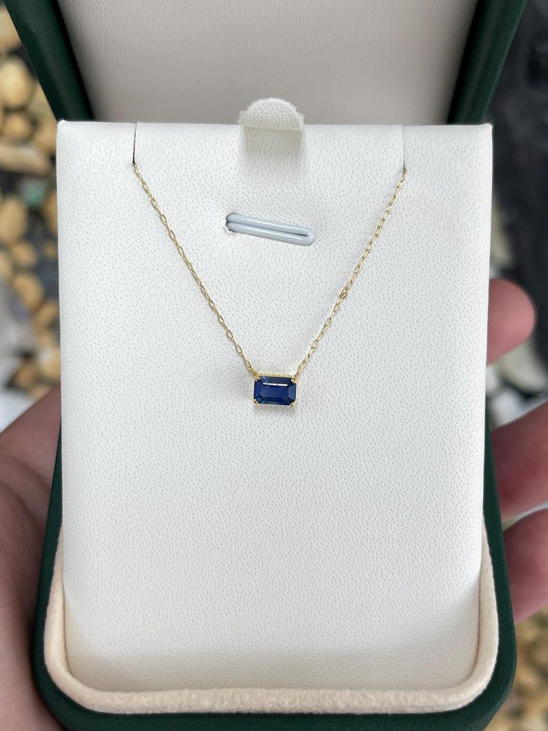 This exquisite solitaire sapphire necklace features a 0.65-carat, emerald-cut sapphire set in an elegant east-west orientation within a secure four-prong setting. The deep blue sapphire is showcased on a delicate 18