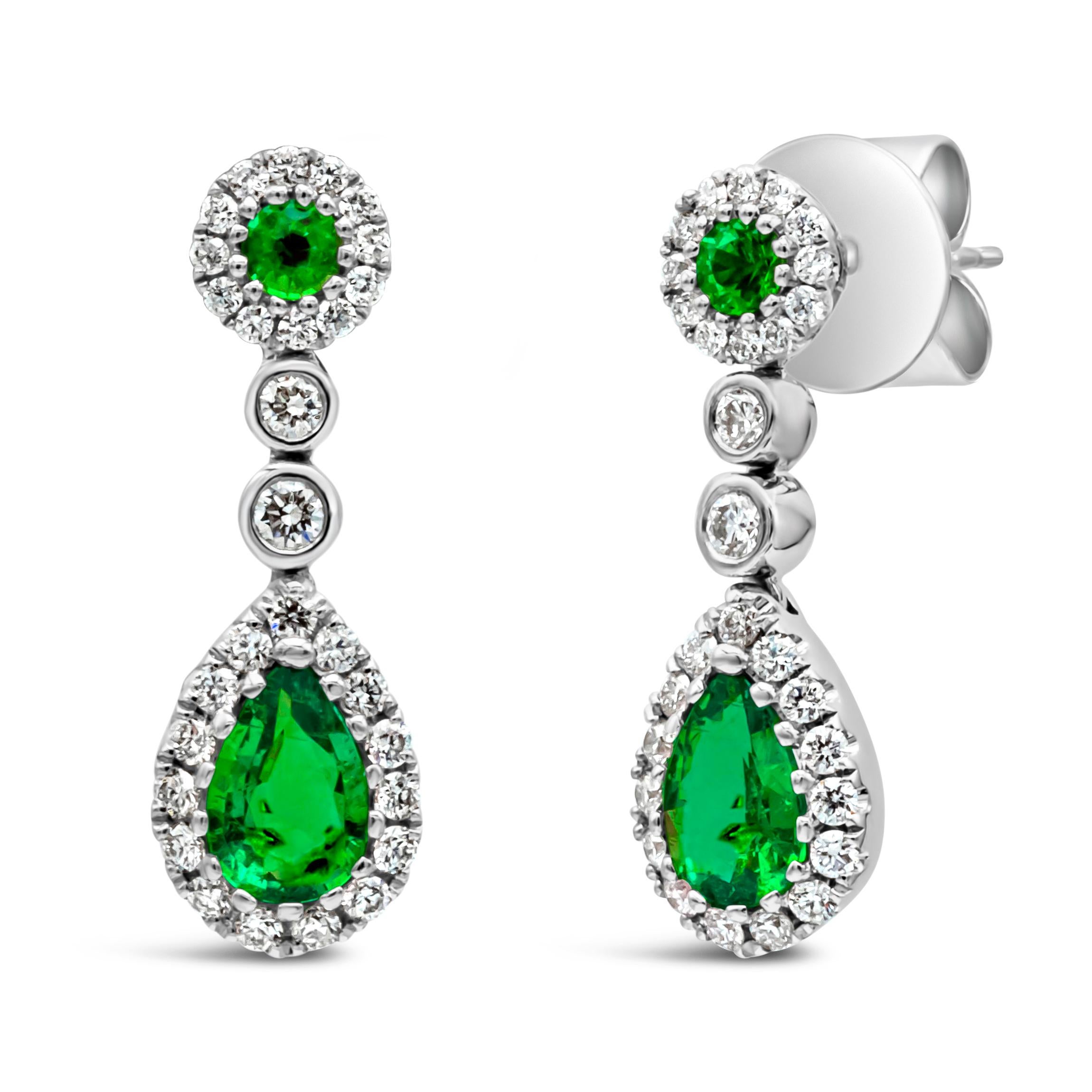 A beautiful and vibrant dangle earrings showcasing a color-rich pear shape Colombian green emerald elegantly set in a halo design. Suspended on a brilliant round shape green emerald halo and spaced with a bezel set round diamond. Colombian green