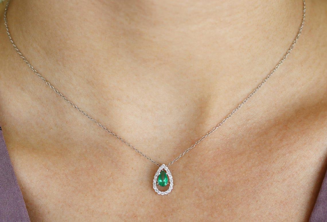 Gezeigt wird ein moderner tropfenförmiger Anhänger mit Smaragd und Diamant. Ein smaragdgrüner Tropfen sitzt bündig in einer sechszackigen Fassung. Dieser Smaragd wurde vollständig von Hand gefertigt und ist in einen Diamanttropfen eingefasst. Der