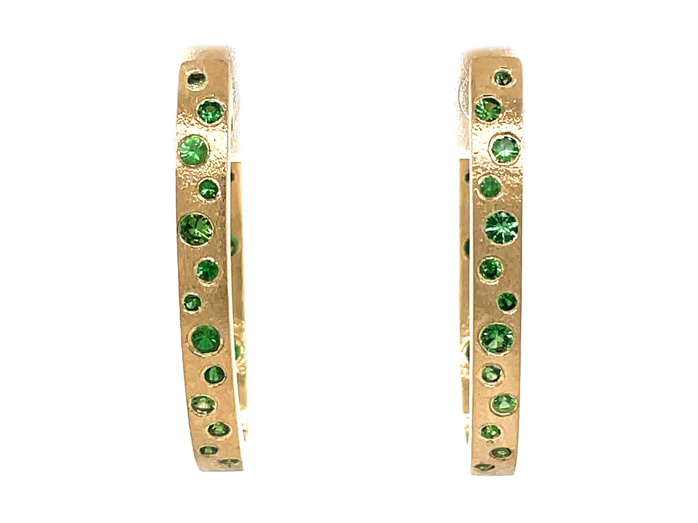 These Green Tsavorite earrings are a stunning and timeless accessory that can add a touch of glamour and sophistication to any outfit. These Flush Set Oval Hoop Earrings contain 0.73 Carats of Round Green Tsavorites , set in 14K Yellow Gold, with a