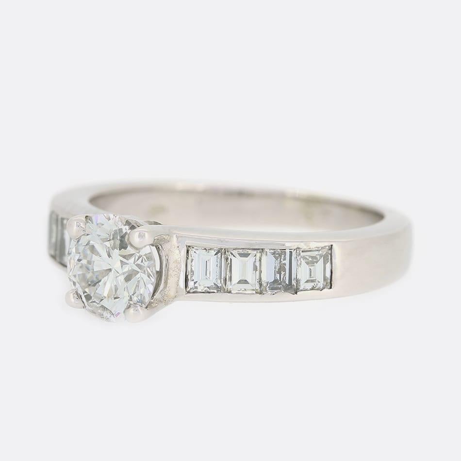 Dies ist ein Solitär-Diamantring aus 18 Karat Weißgold. Im Mittelpunkt des Rings steht ein runder Diamant im Brillantschliff von 0,74 Karat, der in einer erhöhten Fassung mit 4 Klauen gefasst ist. Jede Schulter ist mit 4 Diamanten im Smaragdschliff