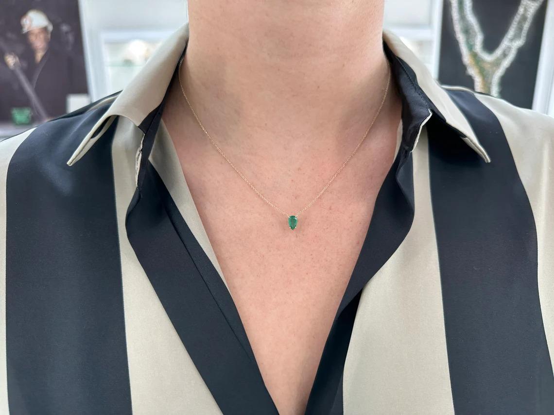 The solitaire pear-cut necklace is a stunning piece of jewelry that features a single emerald stone in a simple four prong setting. The emerald has a medium dark green color that adds to its elegance and beauty. The pendant is attached to a dainty