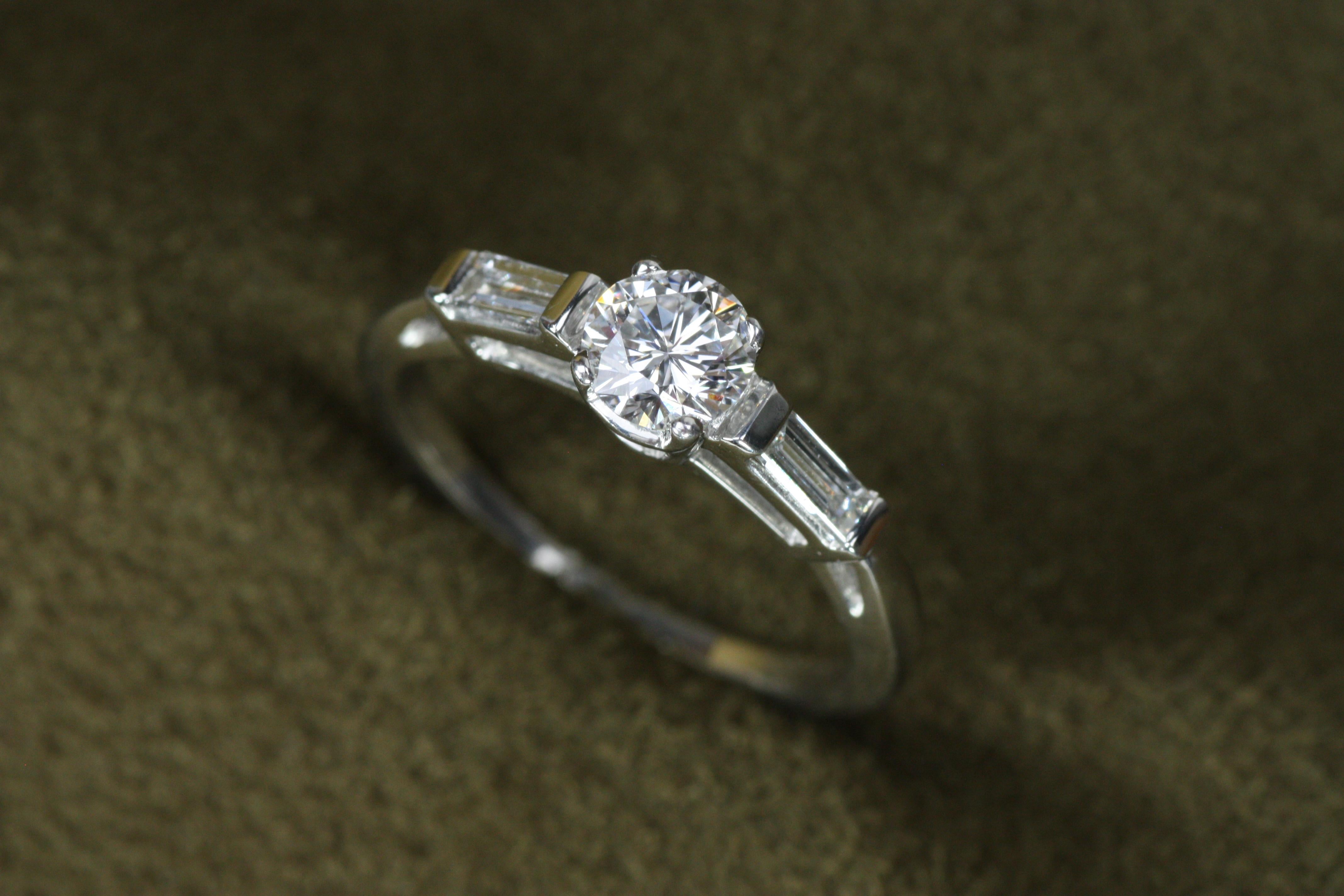 Este moderno anillo de compromiso de tres piedras en platino, inspirado en los años 50, personifica la belleza refinada, con un diamante redondo central de 0,54 ct, certificado por el GIA como 