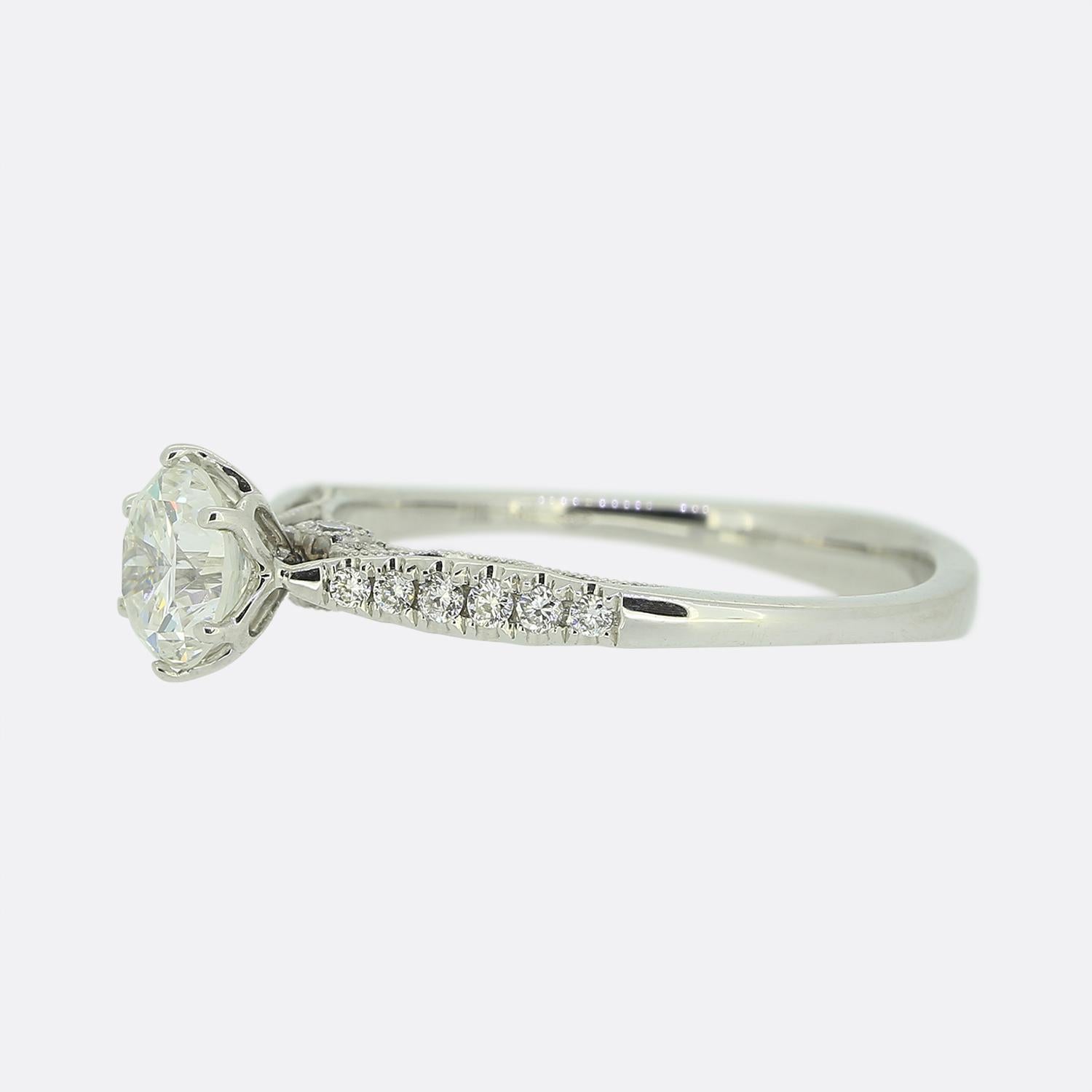 Hier haben wir einen Solitär-Diamantring aus 18 Karat Weißgold. Dieses kunstvolle Stück zeigt eine verschlungene Untergalerie mit einem geschwungenen Perlenmuster, das einen runden Diamanten im Brillantschliff auf jeder Seite beherbergt. In der