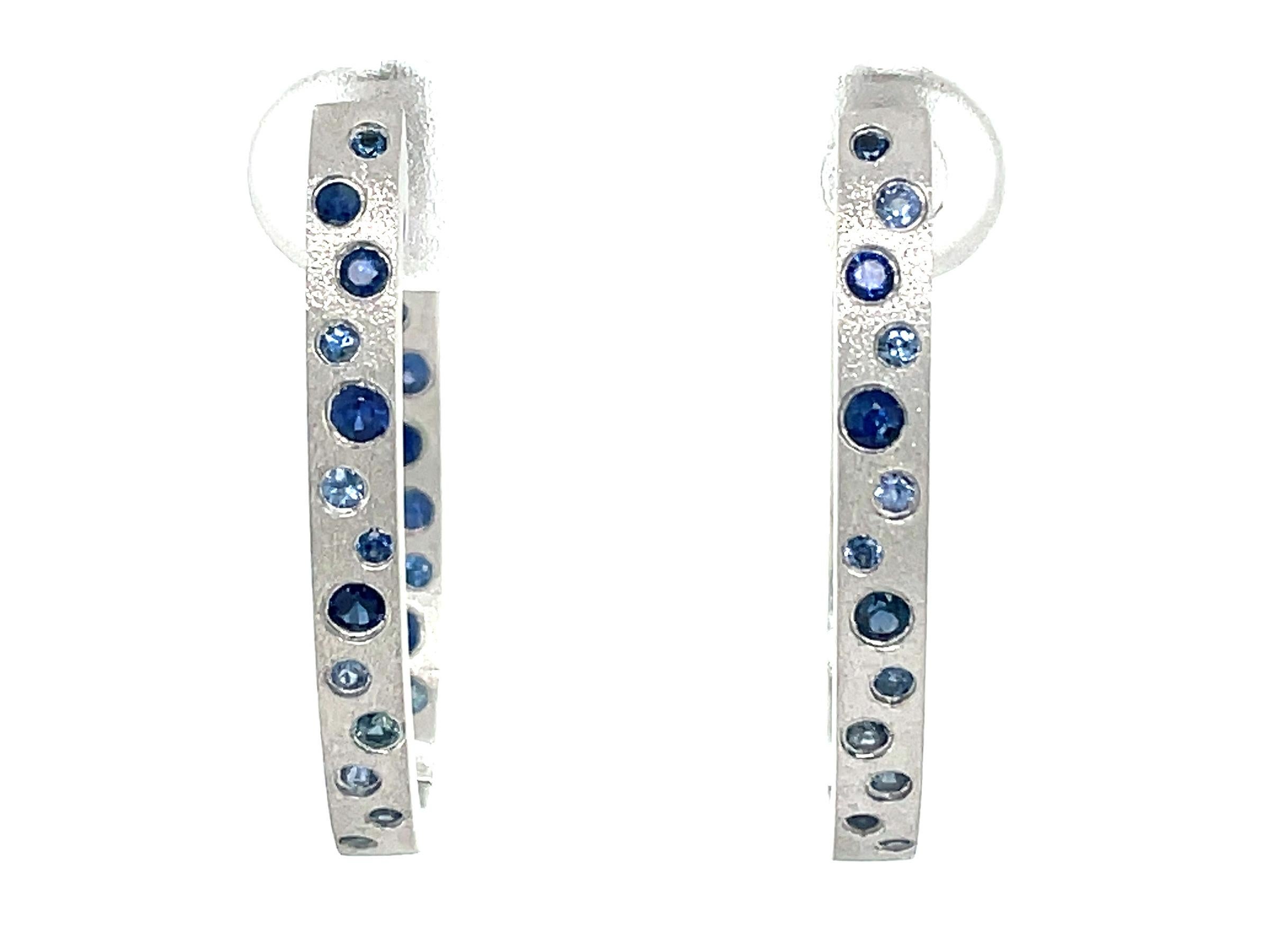 These Sapphire earrings are a stunning and timeless accessory that can add a touch of glamour and sophistication to any outfit. These Flush Set Oval Hoop Earrings contain 0.79 Carats of Round Light, Medium, and Bright Blue Sapphires , set in 14K