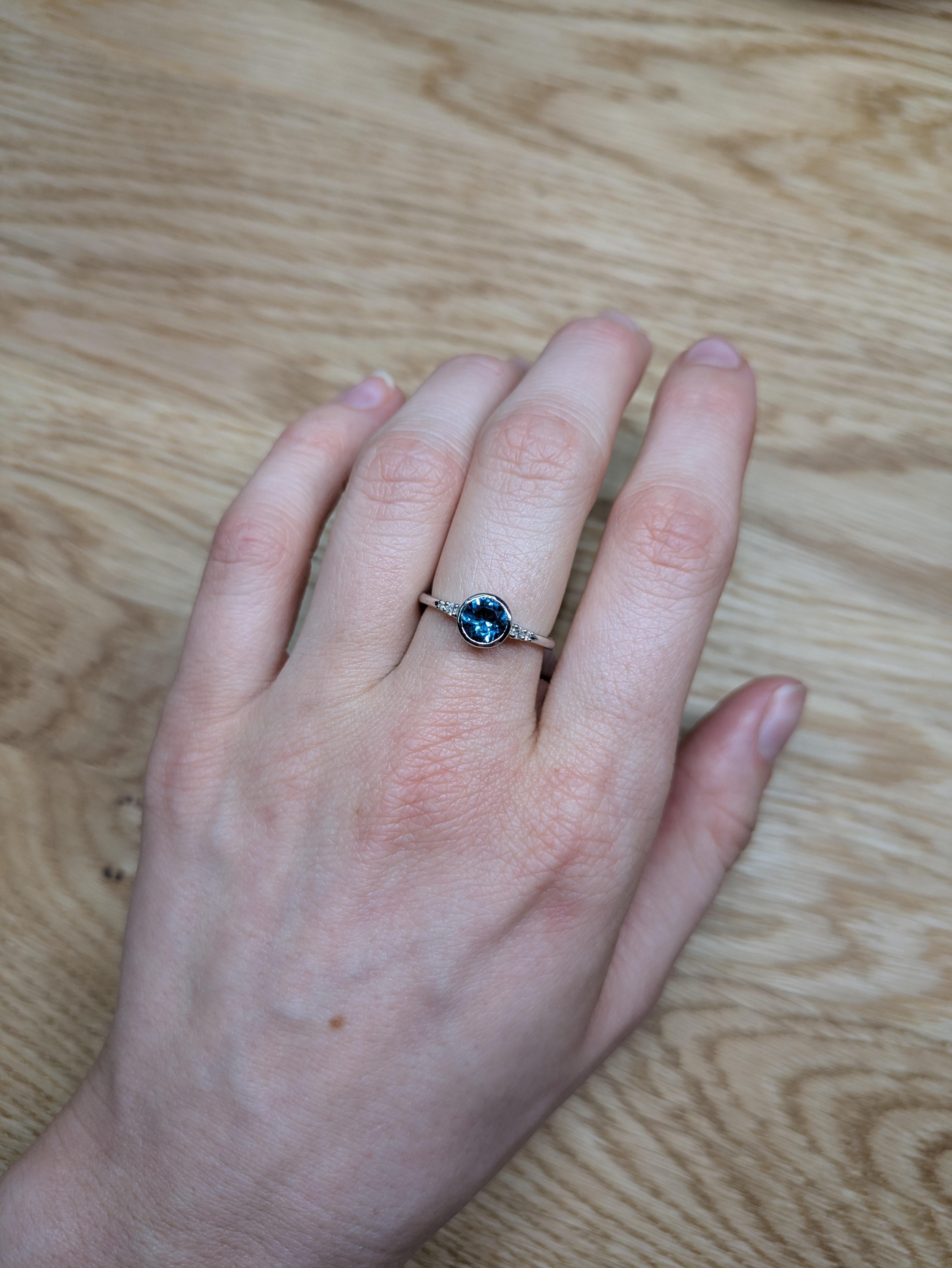 14K White Gold 0.80cts London Blue Topaz Diamond Wedding Ring features a stunning 0.80ct London Blue Topaz in a round 6mm cut, showcasing a deep and captivating blue hue that symbolizes elegance and sophistication. The ring is further enhanced with