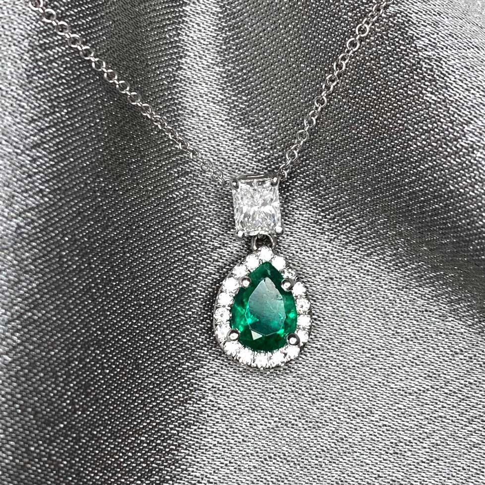 A striking pendant showcasing a pear-shaped emerald of 0.83 carats, cradled in prongs and encircled by a sparkling halo of round brilliant cut diamonds. A radiant cut diamond above the emerald adds a touch of brilliance. The pendant's total diamond