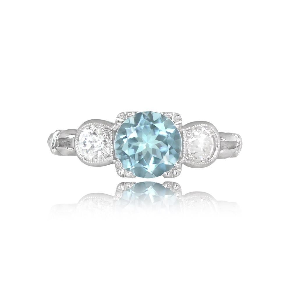 A stunning aquamarine and diamond ring features a 0.86-carat round-cut aquamarine set in prongs, flanked by two bezel-set old European cut diamonds. This exquisite ring is handcrafted in platinum and adorned with intricate hand engravings on the