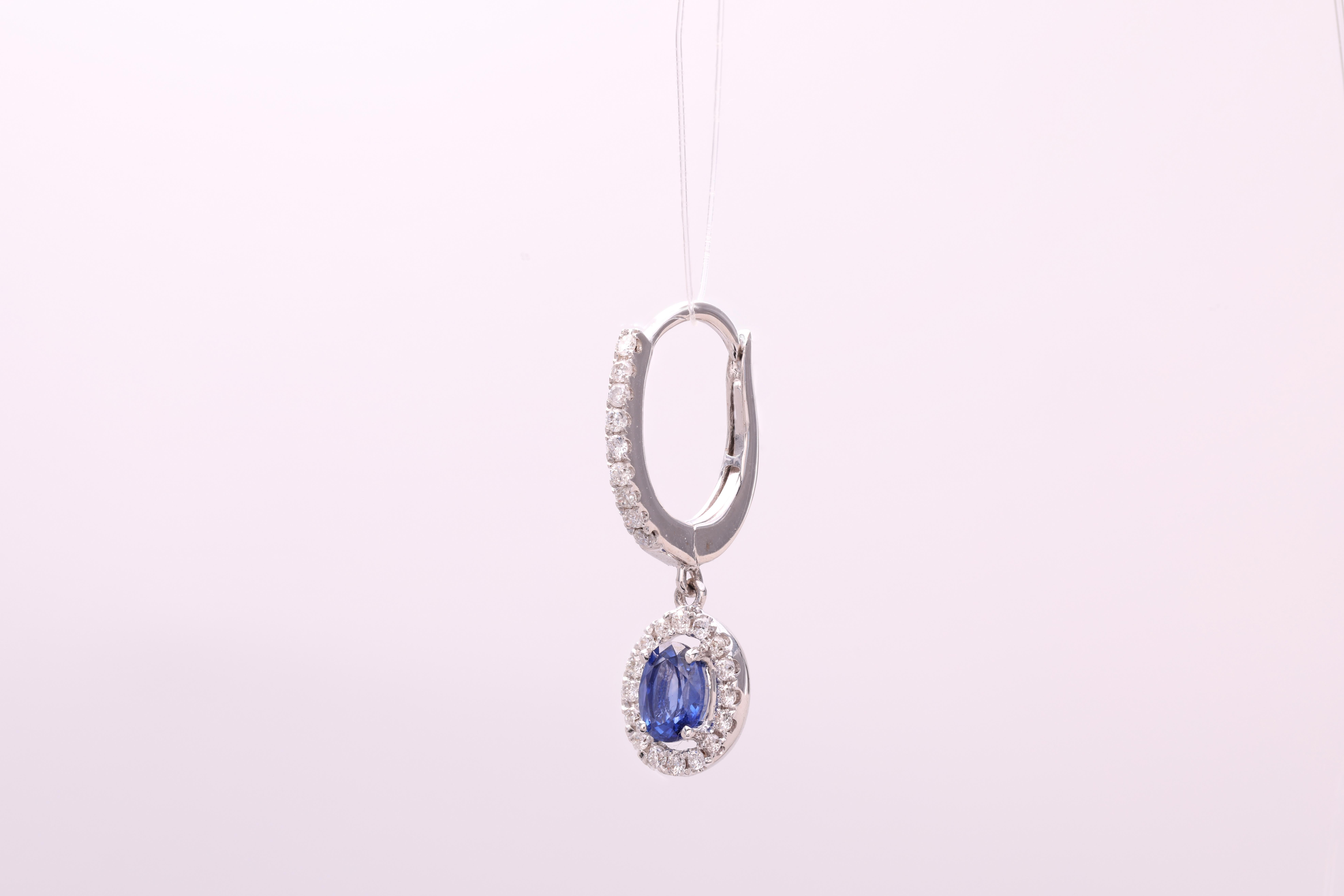 These stunning Halo Drop Sapphire earrings, showcasing an oval sapphire weighing 0.95 carats, elegantly accompanied by 0.33 carats of round, natural diamonds. The sapphire body of the earring dangles delicately from an embellished leverback