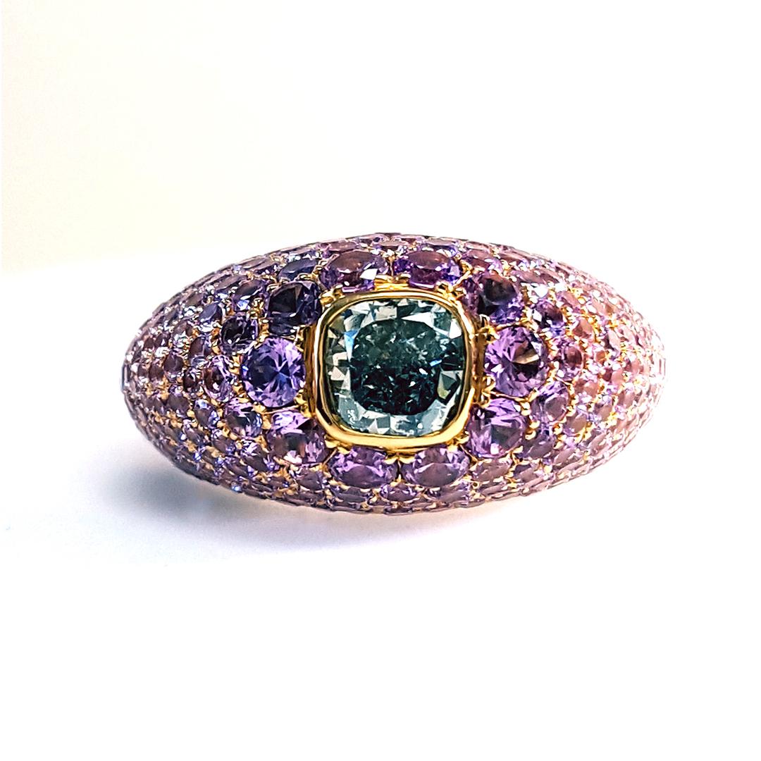 Introducing a one-of-a-kind and exquisite piece: a GIA Certified 1 Carat Fancy Grayish Bluish Green Cushion Cut Diamond & Purple Sapphire Ring. This captivating creation showcases a timeless design that enhances the allure of the central stone,