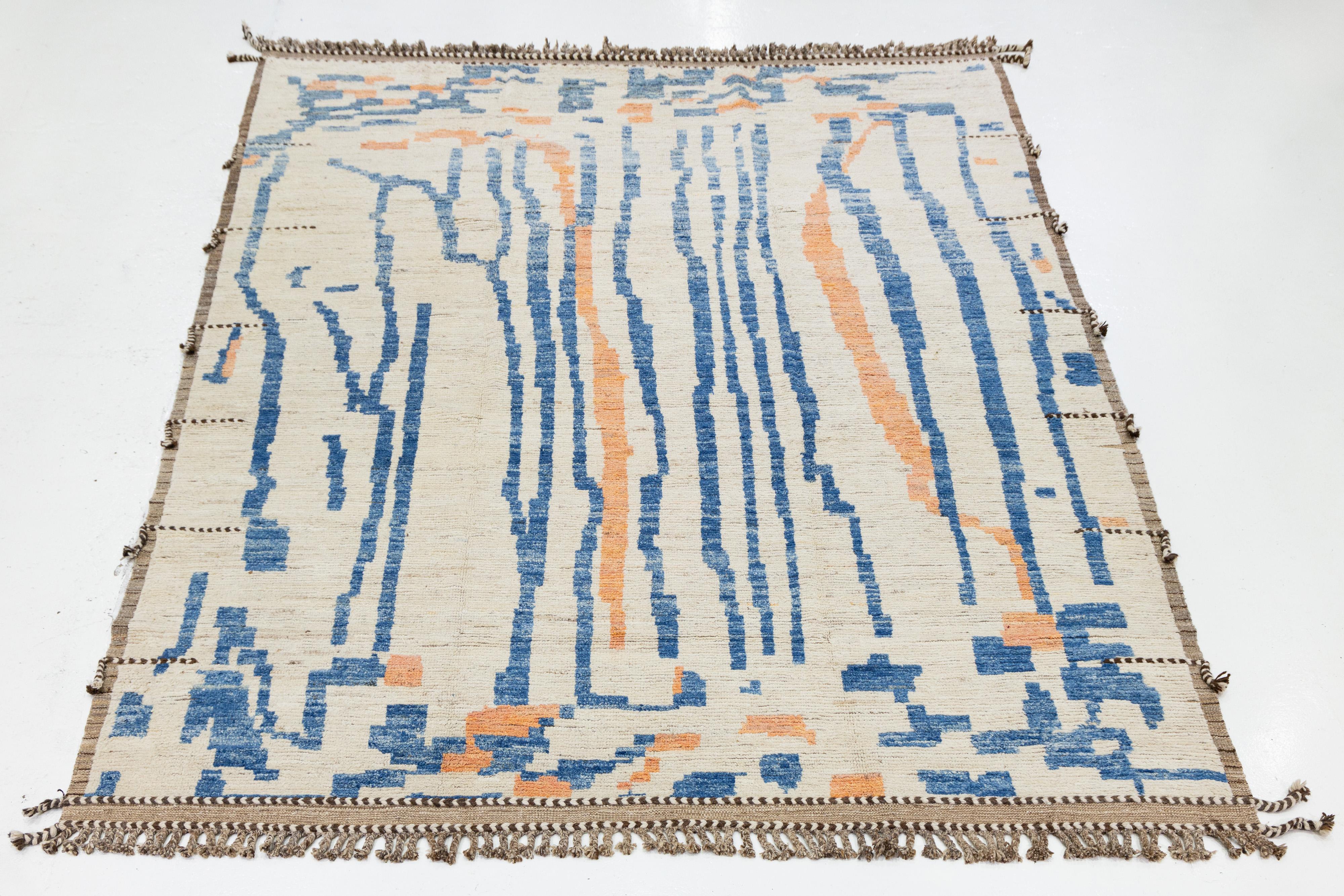 Dieser exquisite, handgefertigte Wollteppich ist ein Beispiel für zeitgenössisches marokkanisches Design mit einem visuell ansprechenden beigen Hintergrund. Das moderne, abstrakte Design, das sich aus einer kühnen Mischung aus Blau, Orange und Braun