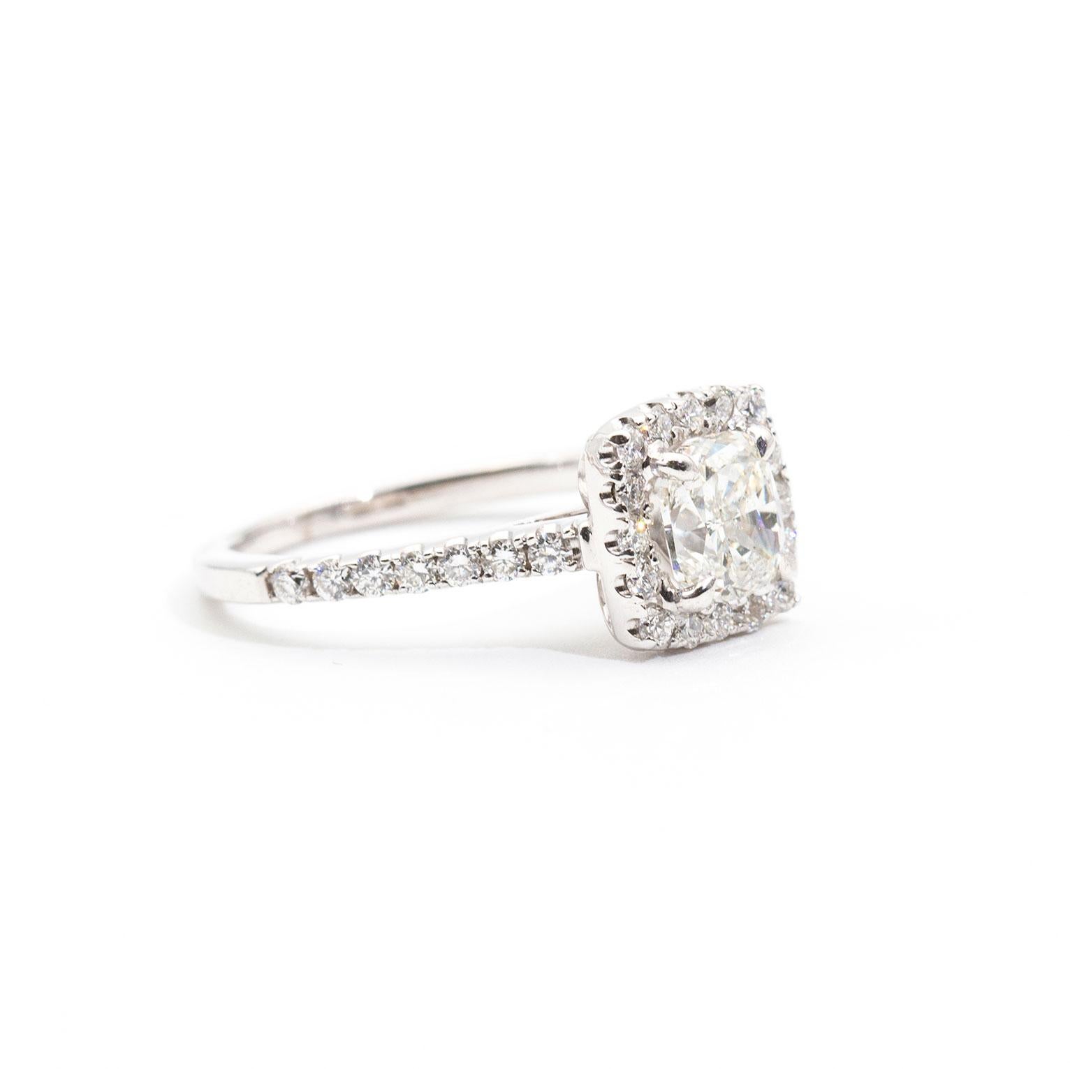 This opulent halo style engagement ring is forged in 18 carat white gold and features a magnificent 1.01 carat cushion modified brilliant cut diamond certified by the Gemological Association of America, GIA. This ring is encrusted with 0.45 carats