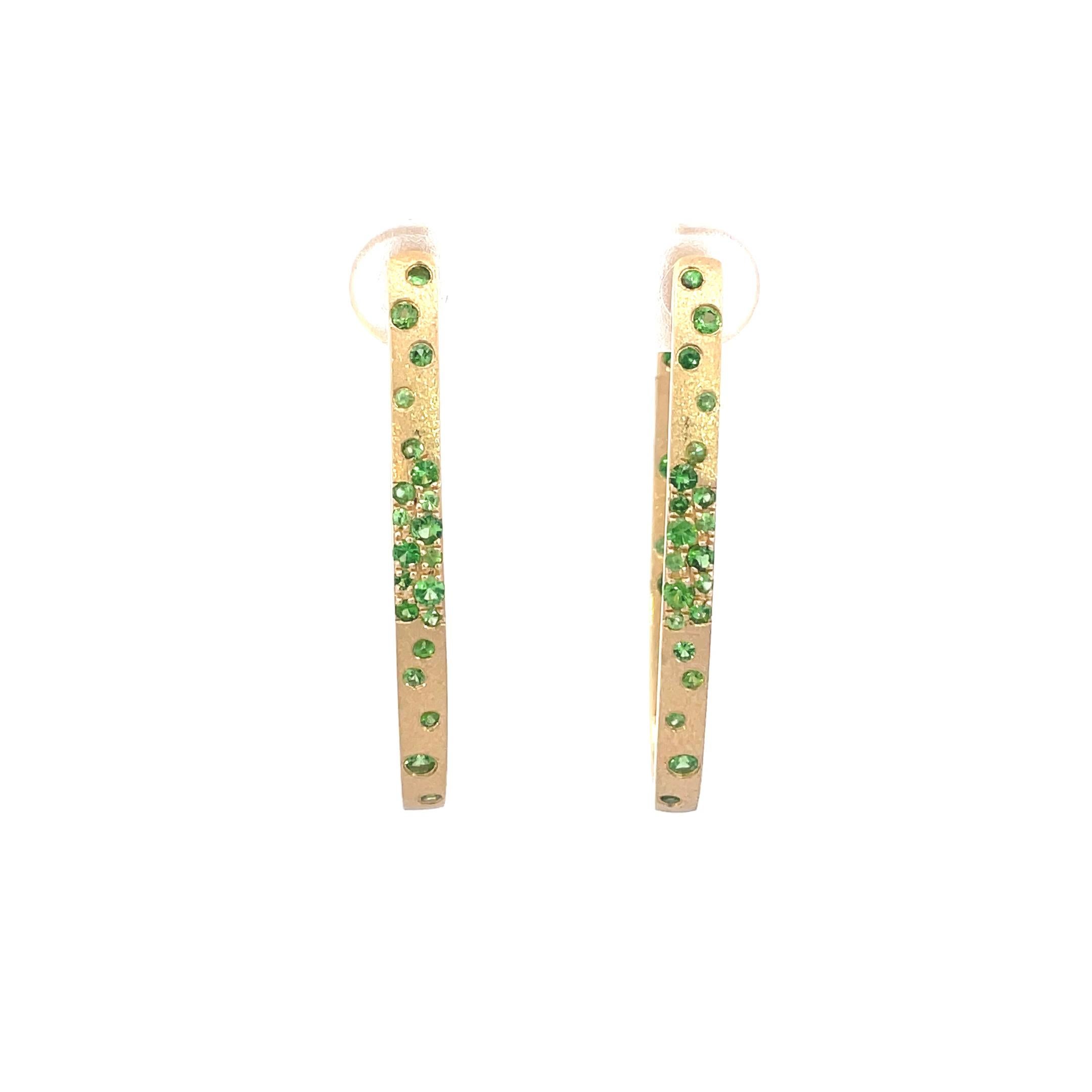 These Tsavorite earrings are a stunning and timeless accessory that can add a touch of glamour and sophistication to any outfit. These Flush Set Oval Hoop Earrings contain 1.01 Carats of Round Green Tsavorites , set in 14K Yellow Gold, with a Satin