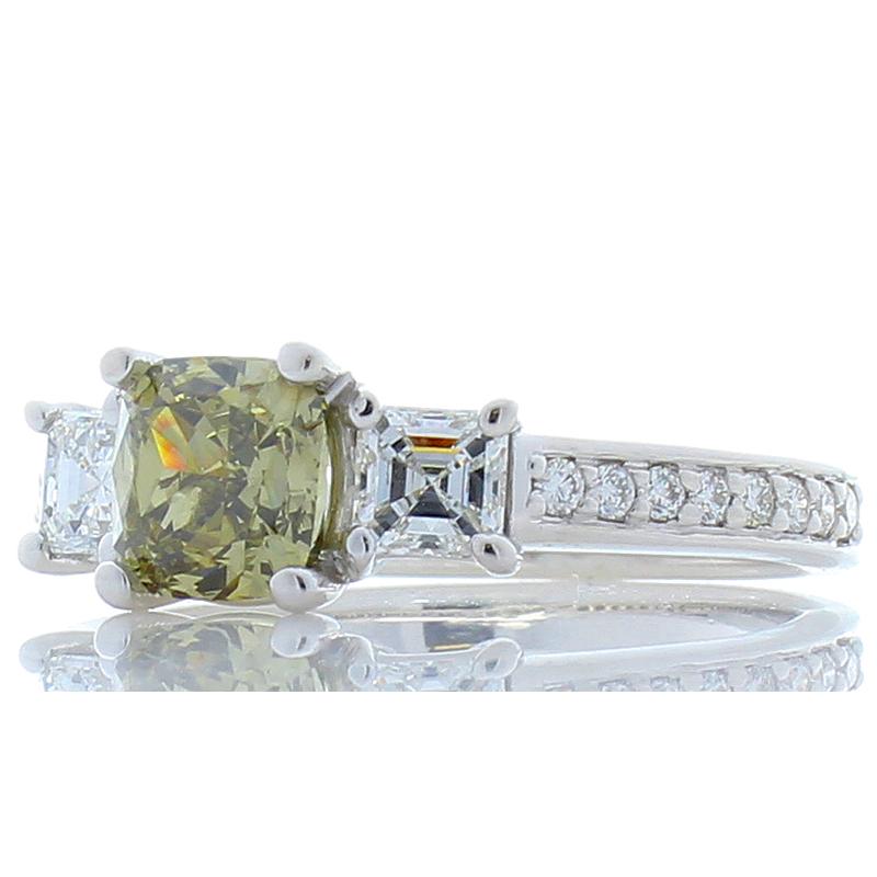 This three stone ring features a 1.02 carat cushion cut, natural fancy greenish brown diamond. It measures 5.7x5.5mm. This natural colored diamond is flanked by two square emerald cut diamonds (F color, VS clarity) totaling 0.48 carats. Round