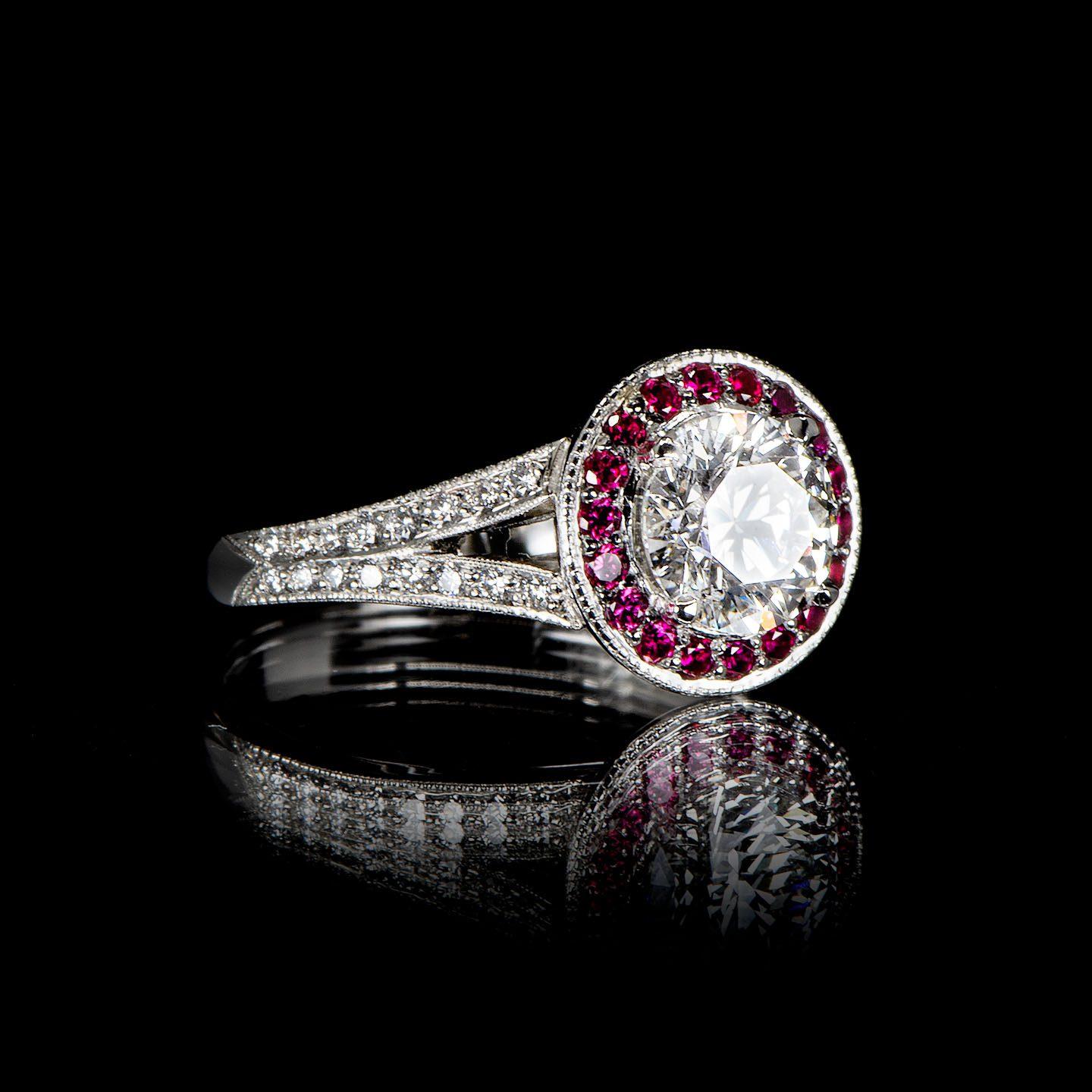 This handcrafted vintage style ring features a 1.02 carat round cut diamond (Colour G, Clarity SI2, laser inscribed, Triple Excellent HRD certificate) surrounded by rubies in a milgrain cluster (halo) setting with black rhodium which sets off their