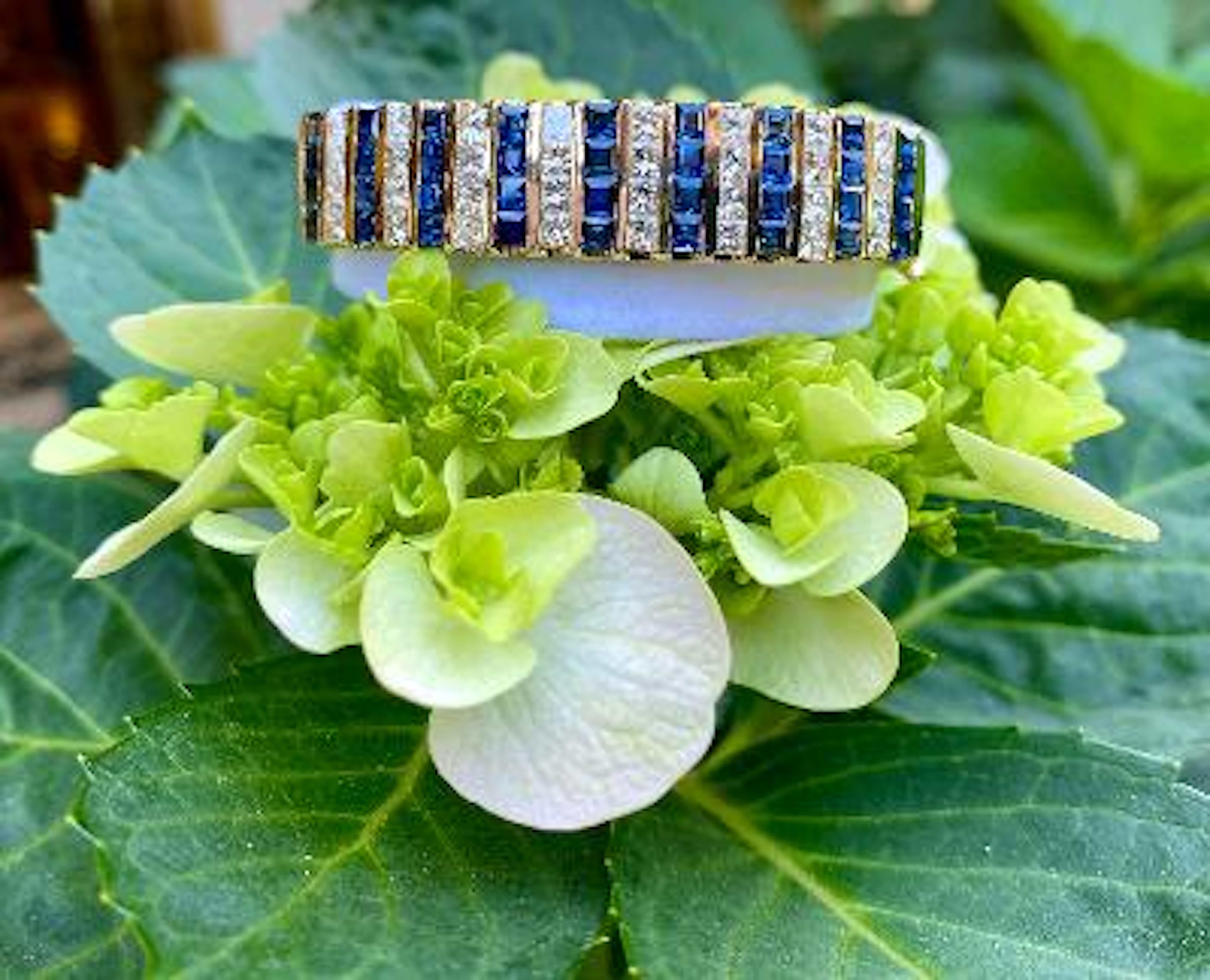 Sparkling diamond and sapphire 14 karat yellow gold estate hinged bangle bracelet features 17 rows of invisibly channel set princess cut white diamonds and square blue sapphires separated between 18 rows of vertical 14 karat gold sections. The total