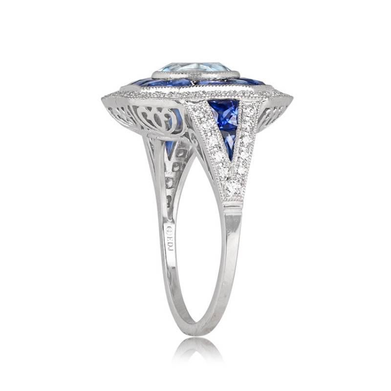 A captivating geometric ring features a round natural aquamarine weighing about 1 carat at its center. The stone is encircled by a double halo of natural French-cut sapphires and round brilliant-cut diamonds. Sapphires and diamonds also adorn the