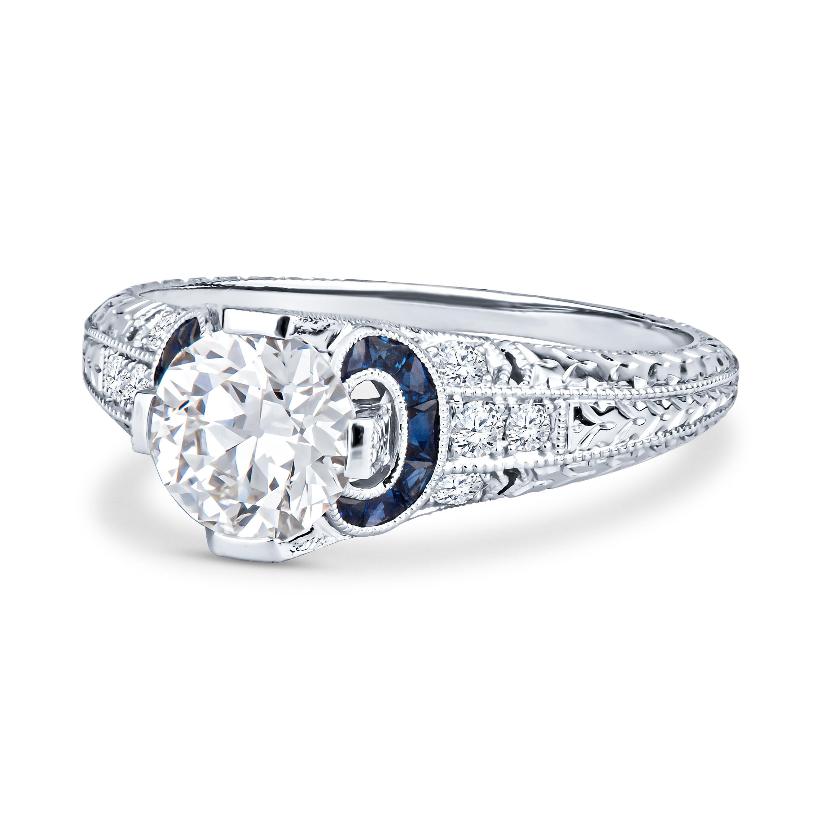 This unique Shaftel Diamonds engagement ring features a 1.03ct round diamond with 0.21ct total weight in blue sapphires and 0.14ct total weight in accent diamonds. The ring is an antique-looking, vintage-inspired engagement ring, with intricate