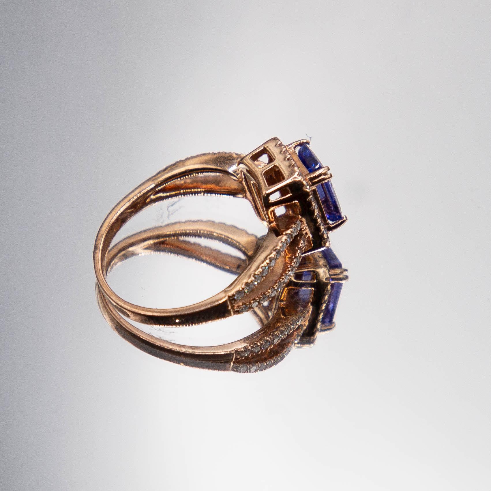 A 14k rose gold ring features a near- flawless emerald-cut Tanzanite at 1.05 carats. The center gem sets in 4 split prongs, radiating a spectacular violetish- blue, resting inside a halo of fine white diamonds. Total diamond weight amongst 68