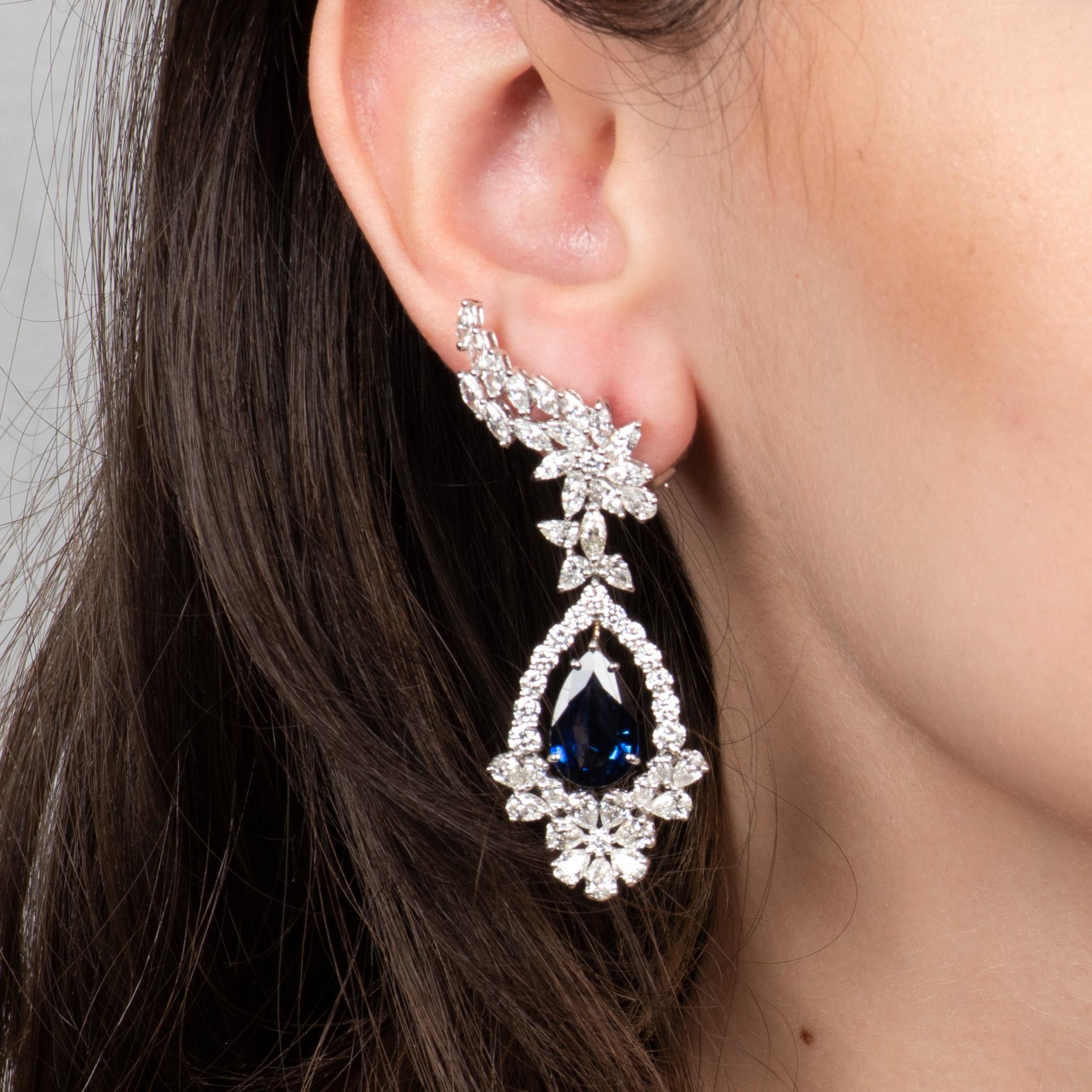 These stunning, extravagant earrings feature a 10.58ctw in a pair of pear shaped natural blue sapphires, surrounded by 10.34ctw in fine white diamonds (in round, marquise, and pear shapes) forming florets and an angel wing pattern on the earlobe.