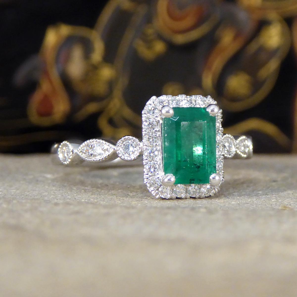 A captivating 1.05ct Emerald Cut Emerald and Diamond Cluster Ring. This ring is a true embodiment of sophistication and grace. Set in the enduring beauty of platinum, this ring features a mesmerising emerald cut emerald at its center with typical