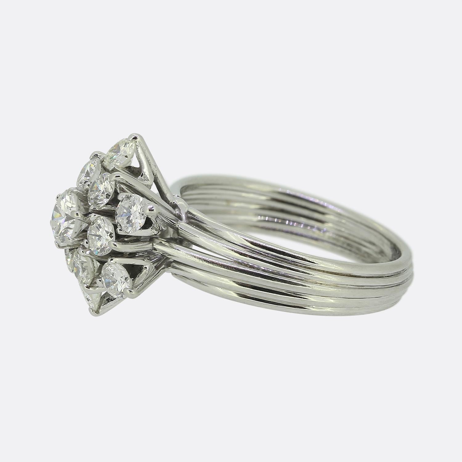 Here we have a stunning diamond cluster ring. A multilayered shank consisting of five slim stacked bands plays host to a single round brilliant cut diamond which sits proud at the centre of the face. This principal diamond is then is surrounded by a
