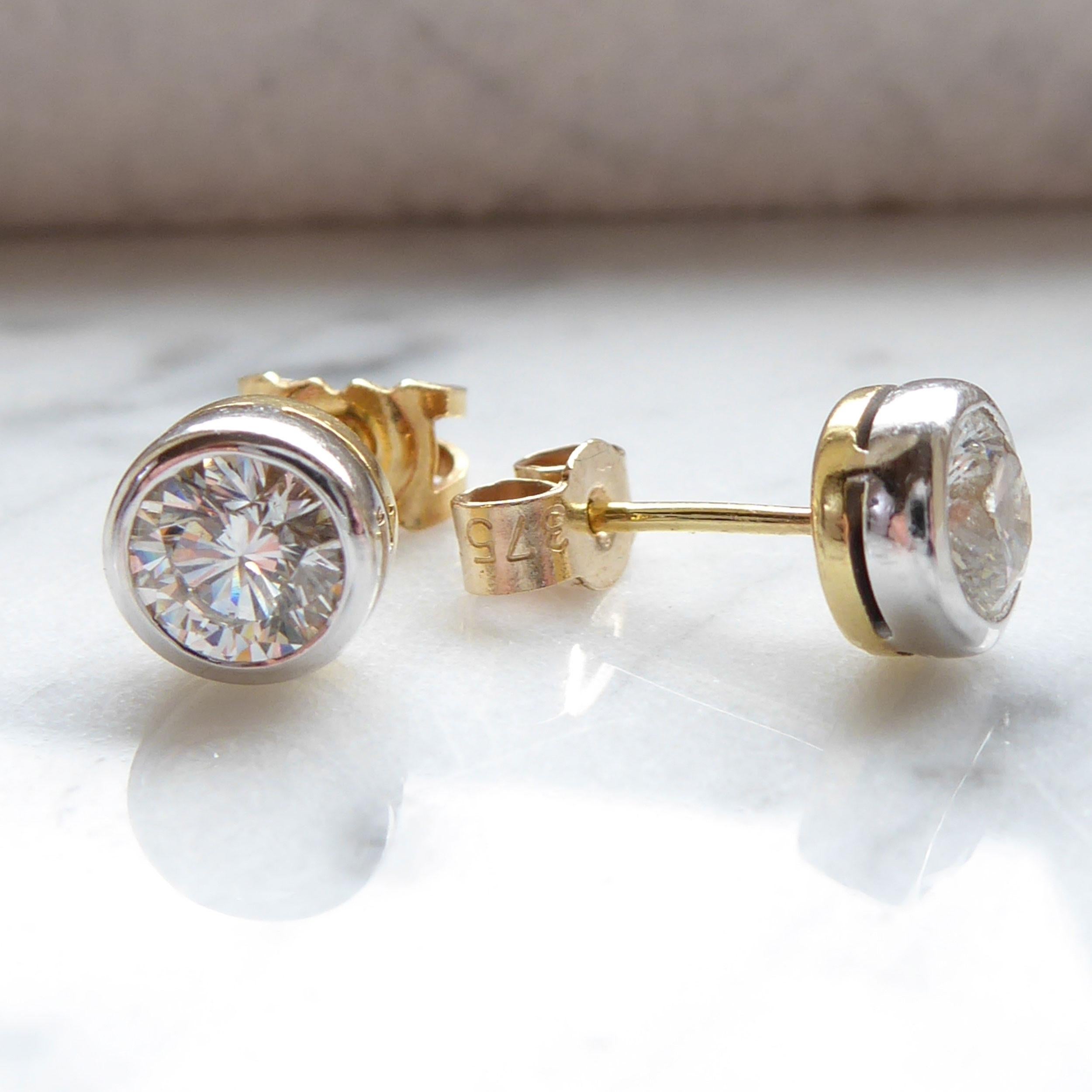 A pair of contemporary diamond stud earrings, each earring set with a round brilliant cut diamond measuring approx. 5.2mm x 3.1mm deep.  Both diamonds are rub-over set to white fronted collet mounts to yellow post and scroll fittings for pierced