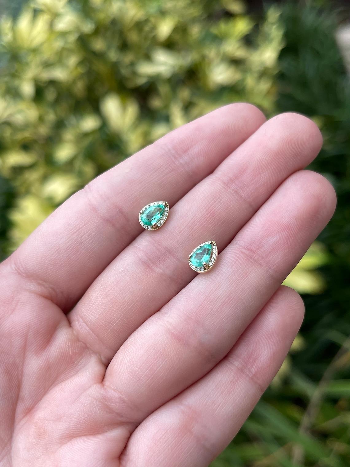 In primo piano c'è una piccola parure di orecchini con smeraldo naturale a forma di pera e diamante VS in oro giallo 14K. Sono esposti smeraldi estratti dalla terra con un'incredibile trasparenza, accentuati da un'aureola di diamanti. L'aureola