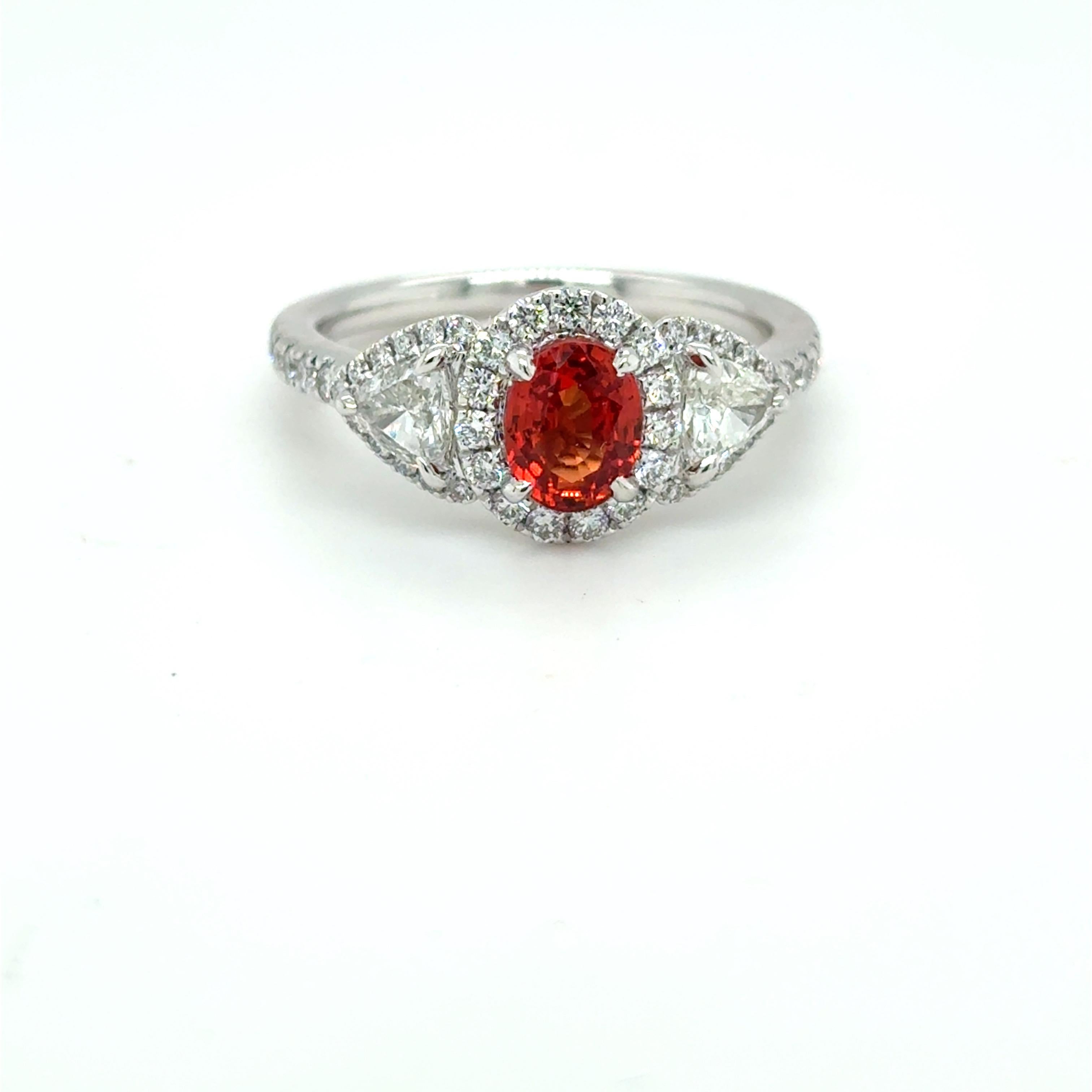 An enchanting 1.08 carat oval Padparadscha sapphire, prized for its delicate blend of pink and orange hues, is the focal point of this elegant platinum ring. Surrounded by a halo of shimmering white diamonds and accented by two 0.48 carat trillion