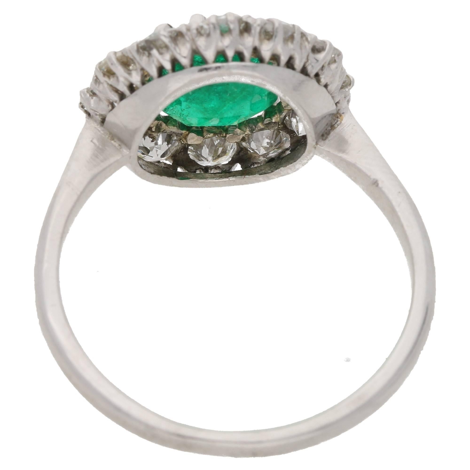 A classic emerald and diamond cluster engagement ring set in platinum. The piece is centrally set with a round faceted emerald, of beautiful colour, secured in twelve platinum claws. The emerald is then haloed with 12 round brilliant cut