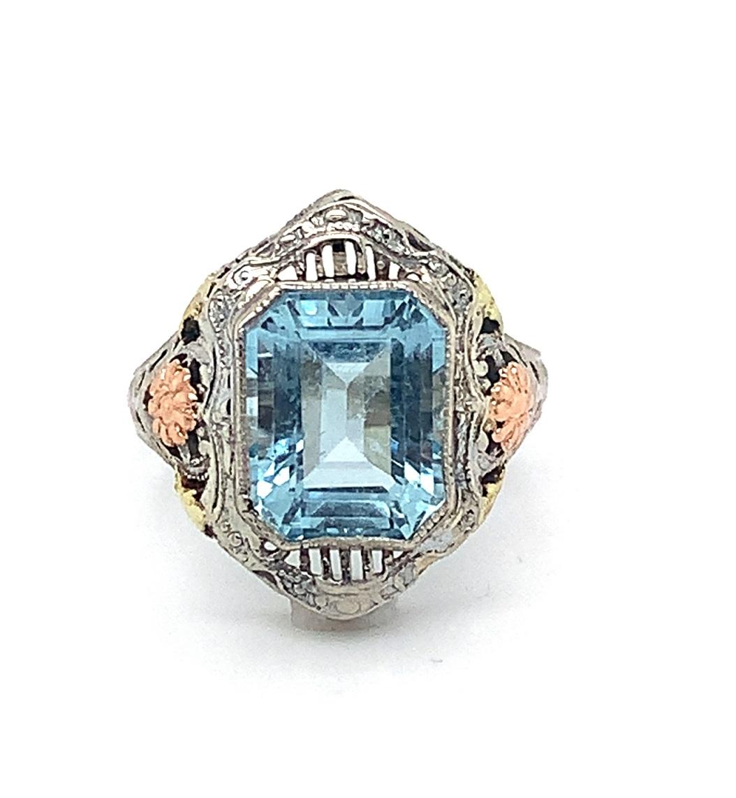 Art Deco 10K white gold filigree ring featuring a beautiful new emerald cut aquamarine weighing 3.28 carats. The light blue aqua measures about 9.5mm x 7.5mm. The gem is accented by applied rose gold flowers with yellow gold leaves. The ring fits a