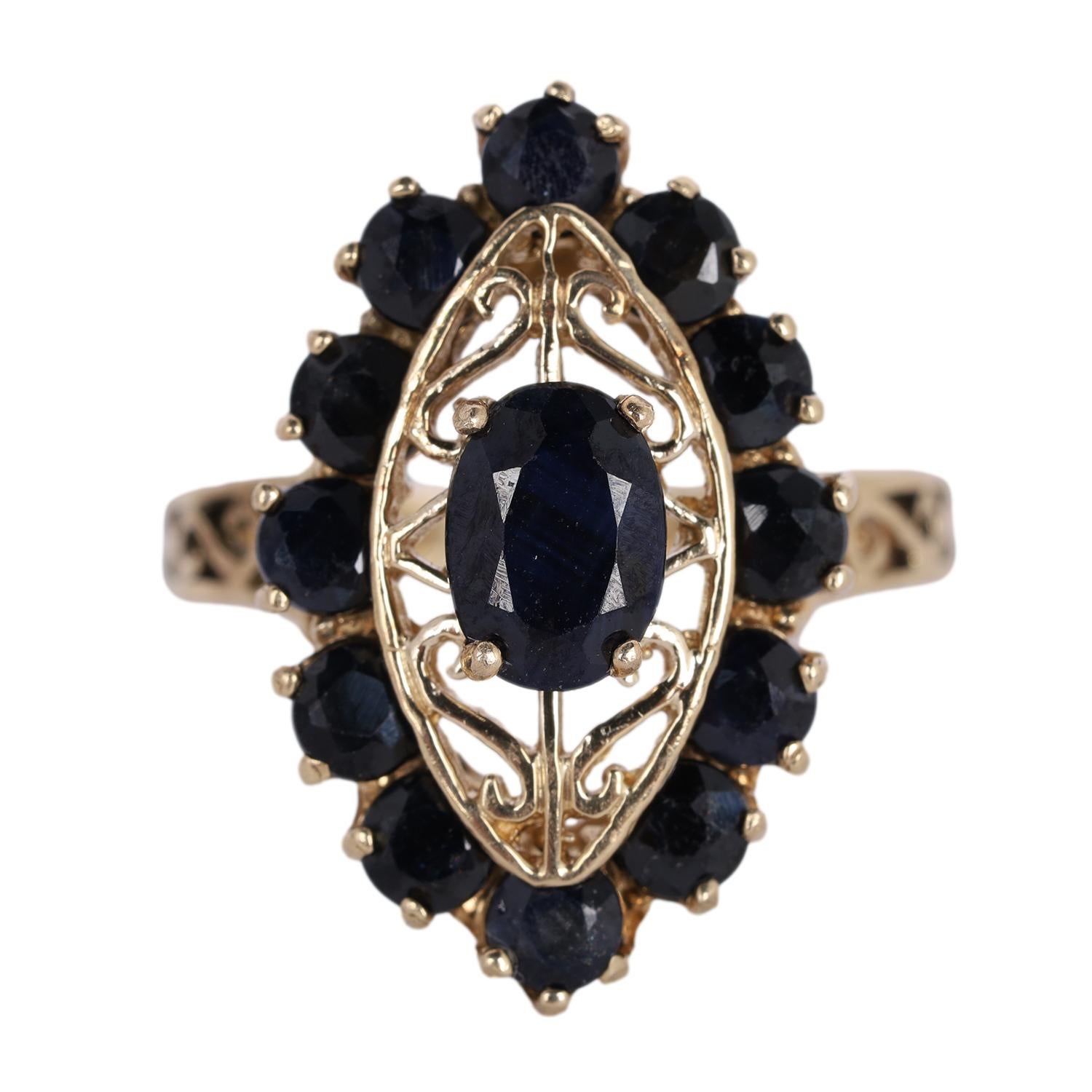The ring features an oval cut and prong set sapphire as the center stone. It measures 7mm in length and 5.02mm in width. Surrounding it in a marquise shaped main setting are 12 additional round cut and prong set sapphires, each measuring