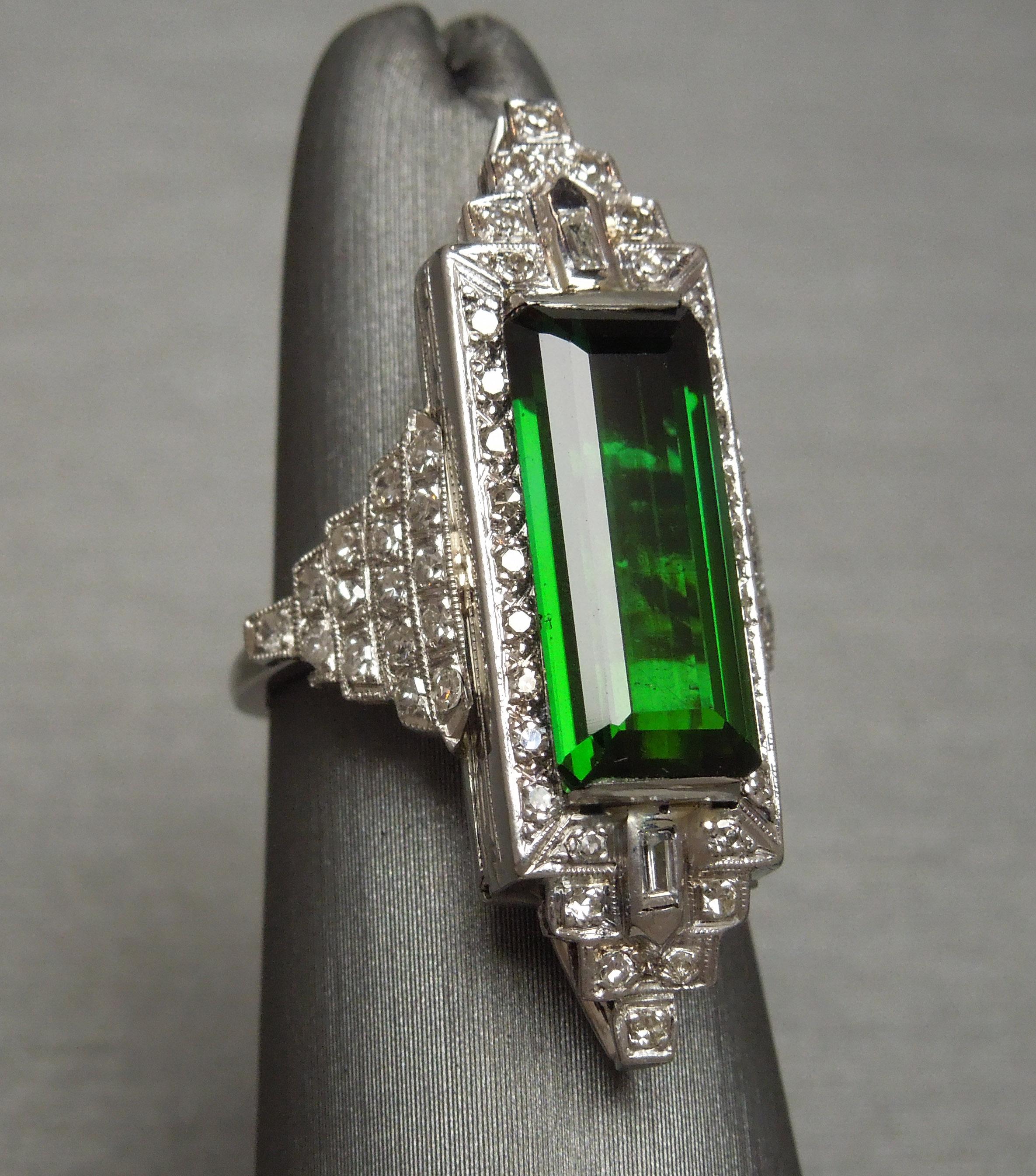 Constructed in the manner of a 1920s Art Deco period ring, this Tourmaline solitaire statement ring features a Central 11 carat Elongated Emerald cut Natural Intense Green Tourmaline at 16.5mm in length x 7.1mm in width. Bordering Tourmaline are a