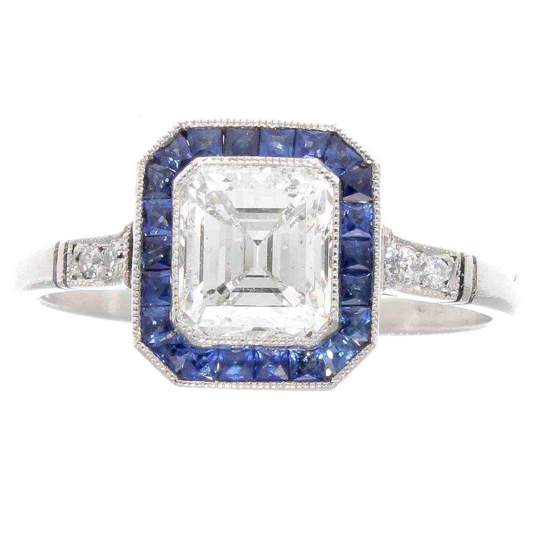 A harmonious creation of tradition, color and design. Featuring a 1.10 carat emerald cut diamond that is I color, SI1 clarity surrounded by a halo of navy blue sapphires calibrated specially to fit around this diamond and in this hand crafted