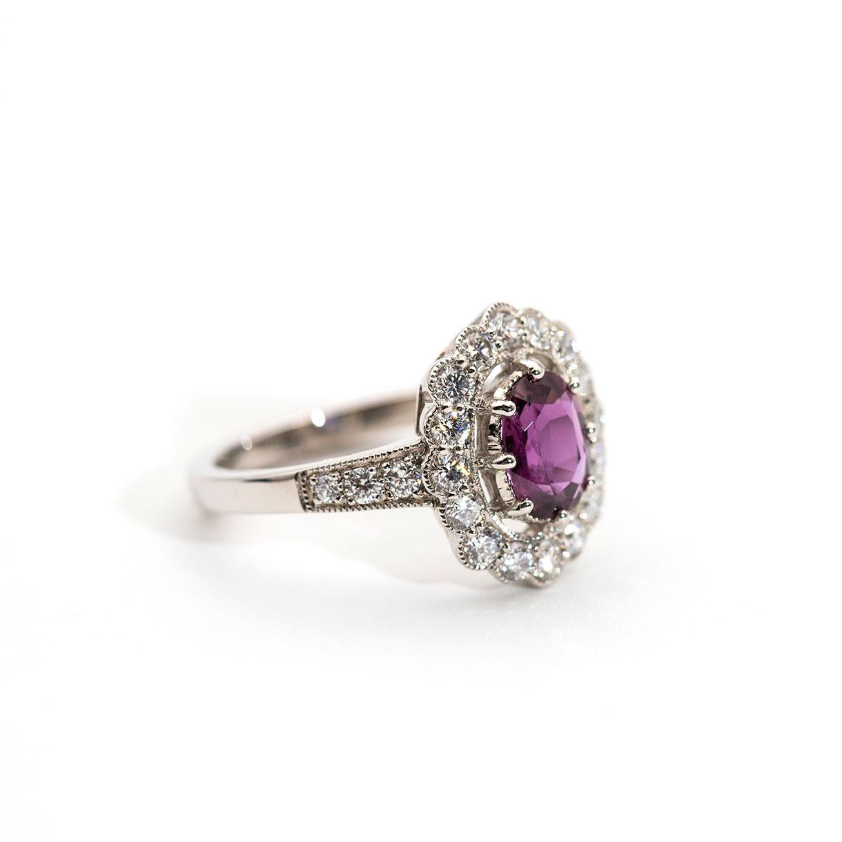 Forged in platinum, this delightful vintage-inspired ring boasts an alluring 1.10 carat bright purple-red faceted natural ruby held in an attractive eight claw setting and surrounded by a striking halo of sixteen shimmering round brilliant cut