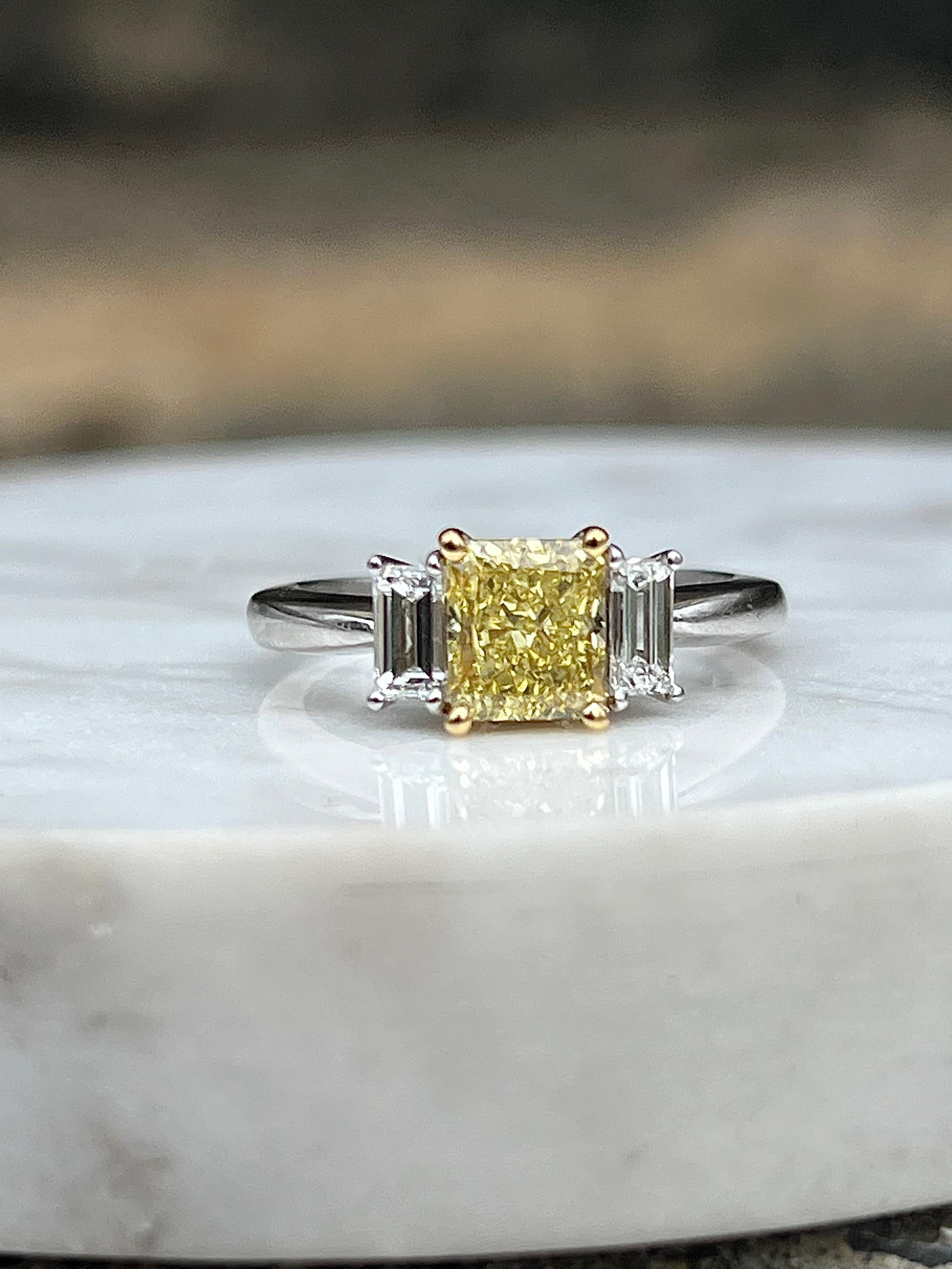 This contemporary three-stone engagement ring elegantly blends platinum and 18 karat yellow gold, creating a timeless setting for a radiant cut fancy yellow diamond of approximately 1.10 carats with VS2 clarity. The vivid yellow center stone is