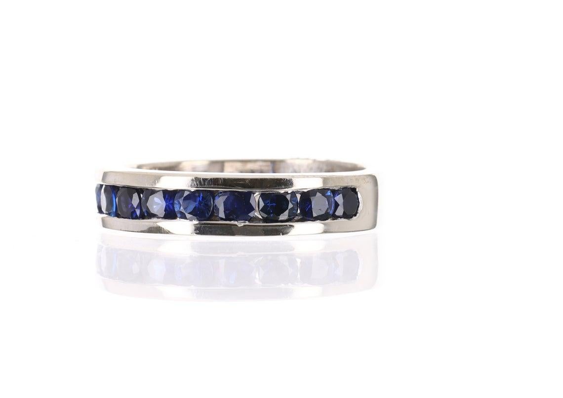 Displayed is a 1.10 TCW blue sapphire band in 14K solid white gold. Soft lines highlight the smooth edges of this ring and provide texture in its simple design. Eleven, bright-blue sapphires are channel set and are of excellent quality. The earth